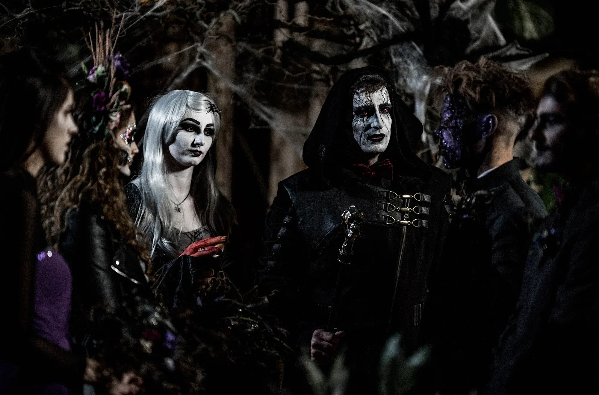Eine düstere Hochzeitsszene mit einer Gruppe von Gästen in Halloween-inspirierter Schminke und Gothic-Kleidung. Die Atmosphäre ist dunkel und mystisch.