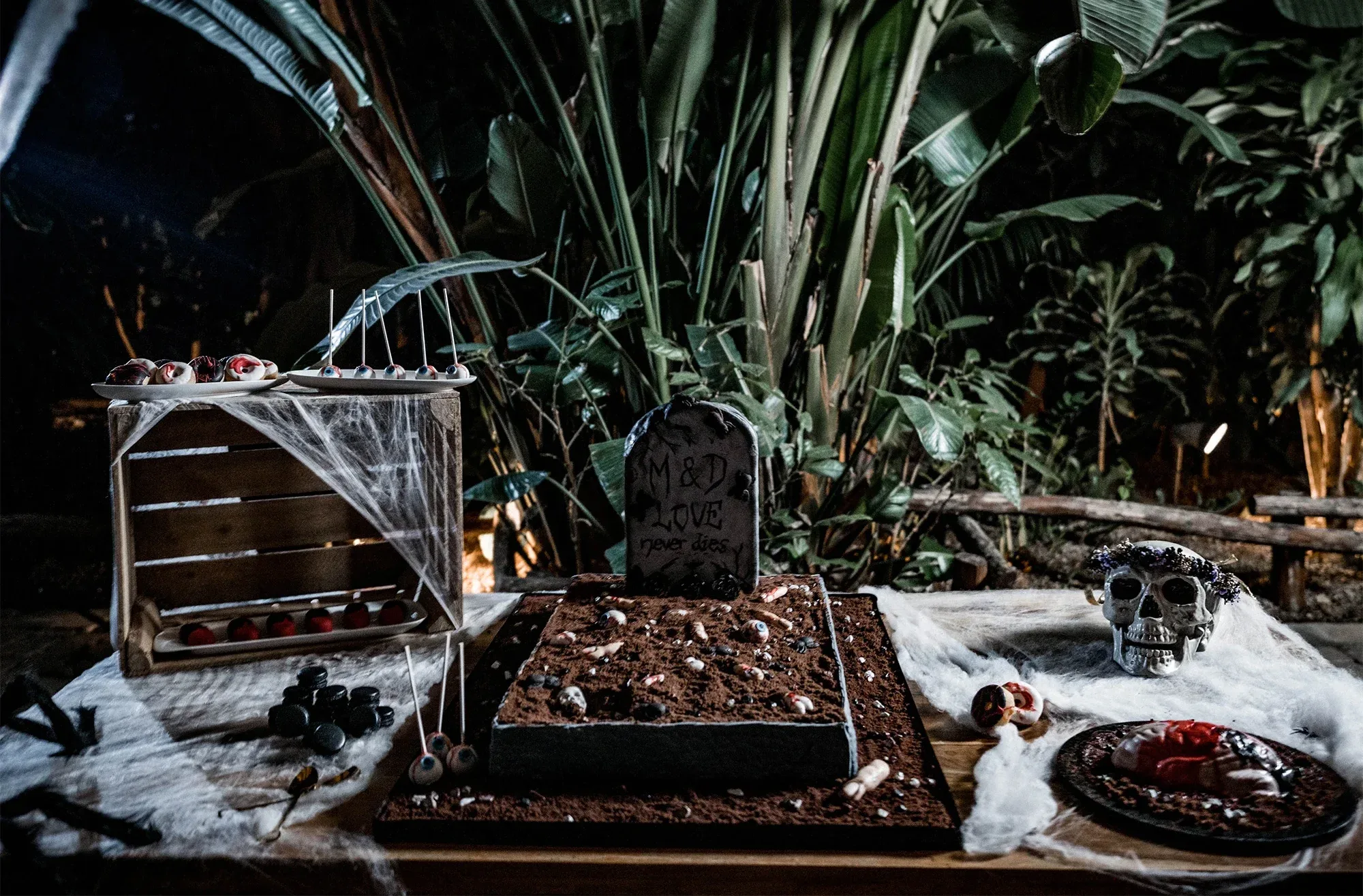 Ein gruseliger Sweet Table mit Grabstein-Deko, Spinnweben und Totenkopf-Dekor, umgeben von tropischen Pflanzen. Ein Halloween-inspirierter Hochzeitstisch.
