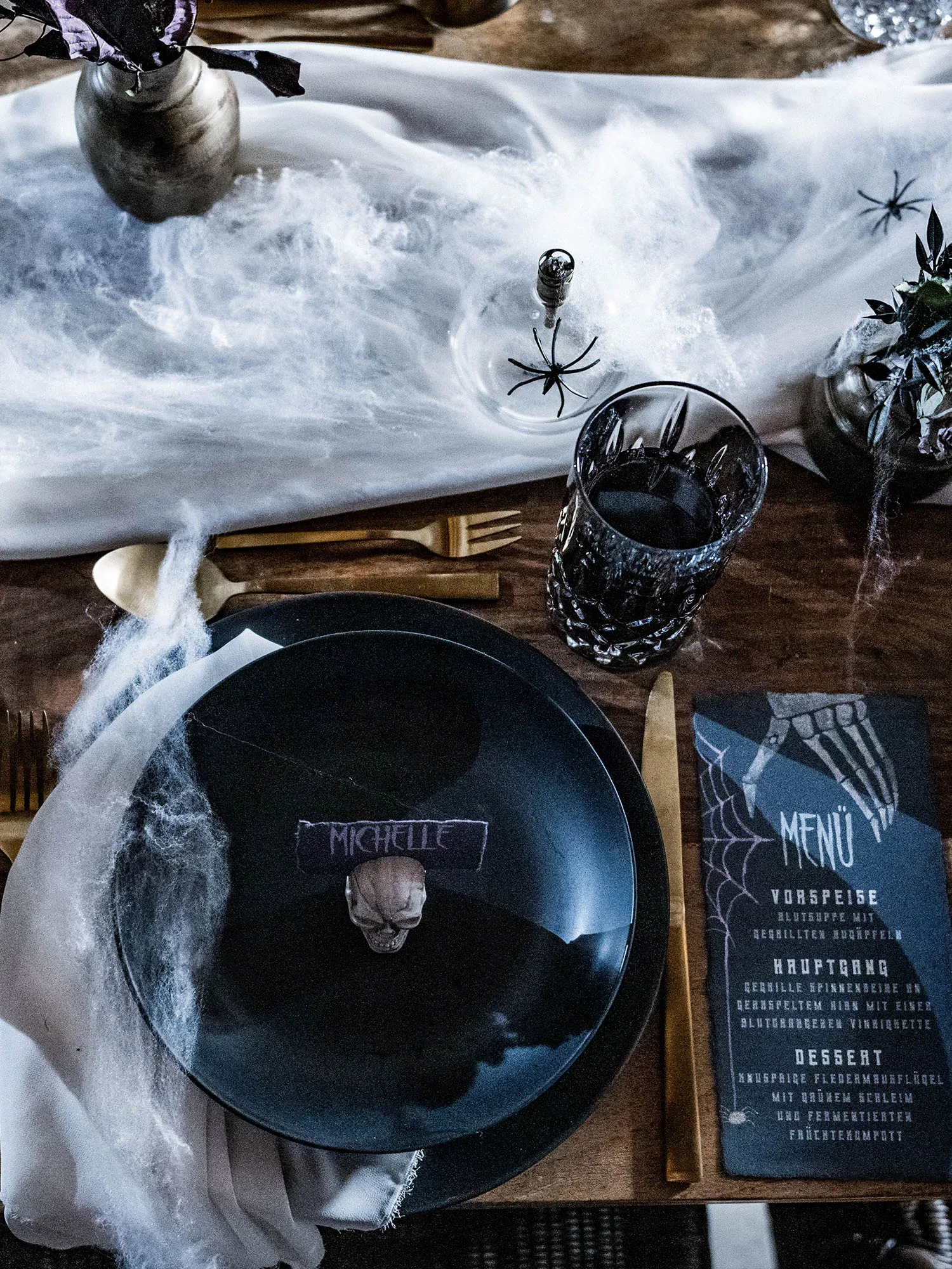 Ein gruseliges Tischgedeck mit schwarzem Teller, Totenkopf-Namensschild und Menükarte im Halloween-Stil, dekoriert mit Spinnweben und schwarzem Glas.