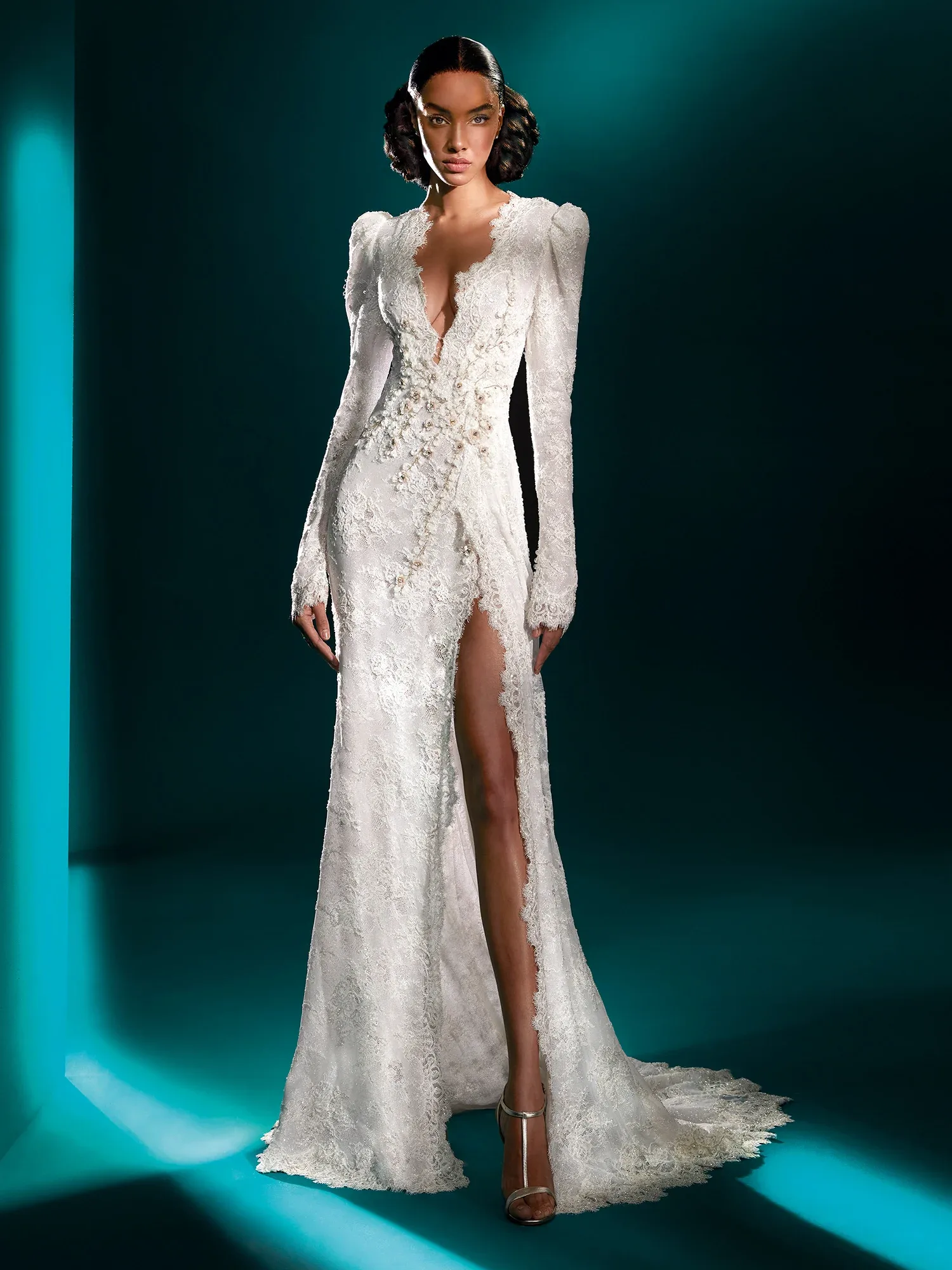 Vorderansicht eines tief ausgeschnittenen Brautkleides mit langen Spitzenärmeln und einem hohen Beinschlitz. Die detaillierten floralen Verzierungen und der figurbetonte Schnitt verleihen dem Kleid eine dramatische Eleganz.