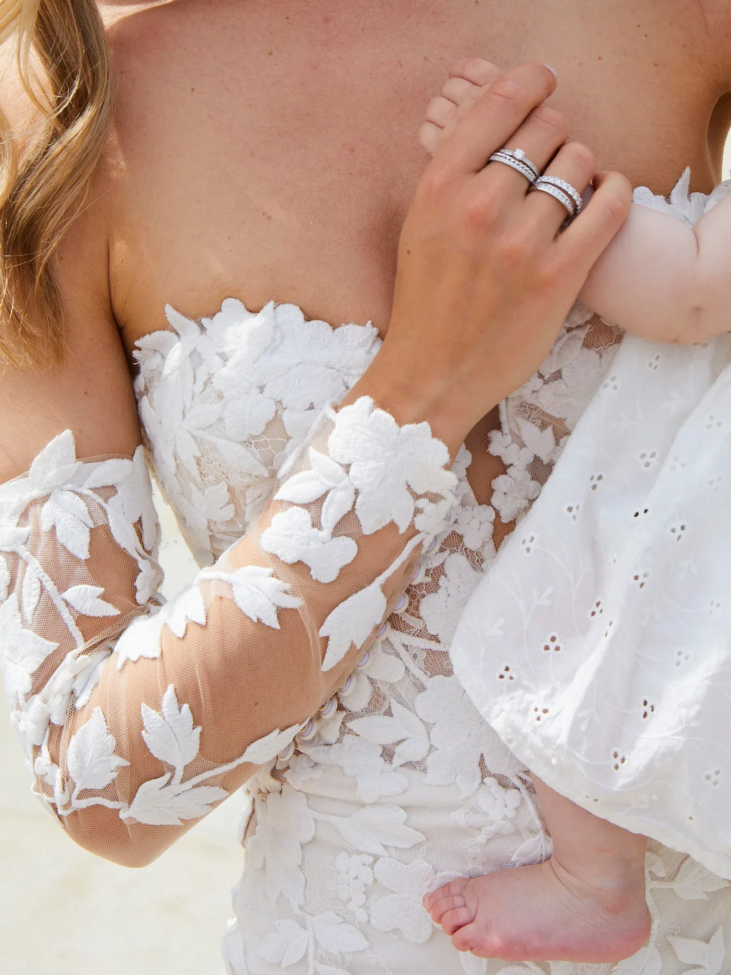 Nahaufnahme einer Braut, die ein Baby im Arm hält. Die Braut trägt ein Kleid mit floraler Spitze und zeigt ihren Ehering, während das Baby sanft ihre Hand hält.