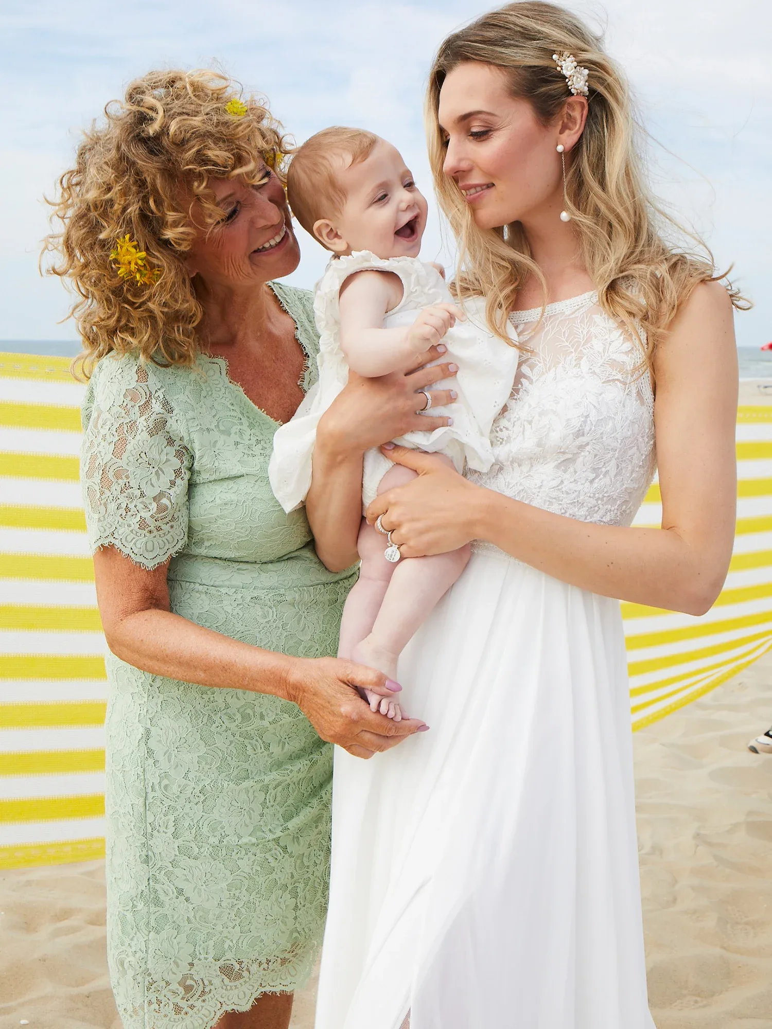 Drei Generationen am Strand: Eine Großmutter in einem mintfarbenen Kleid, eine Braut im weißen Kleid und ein lachendes Baby auf dem Arm der Braut.