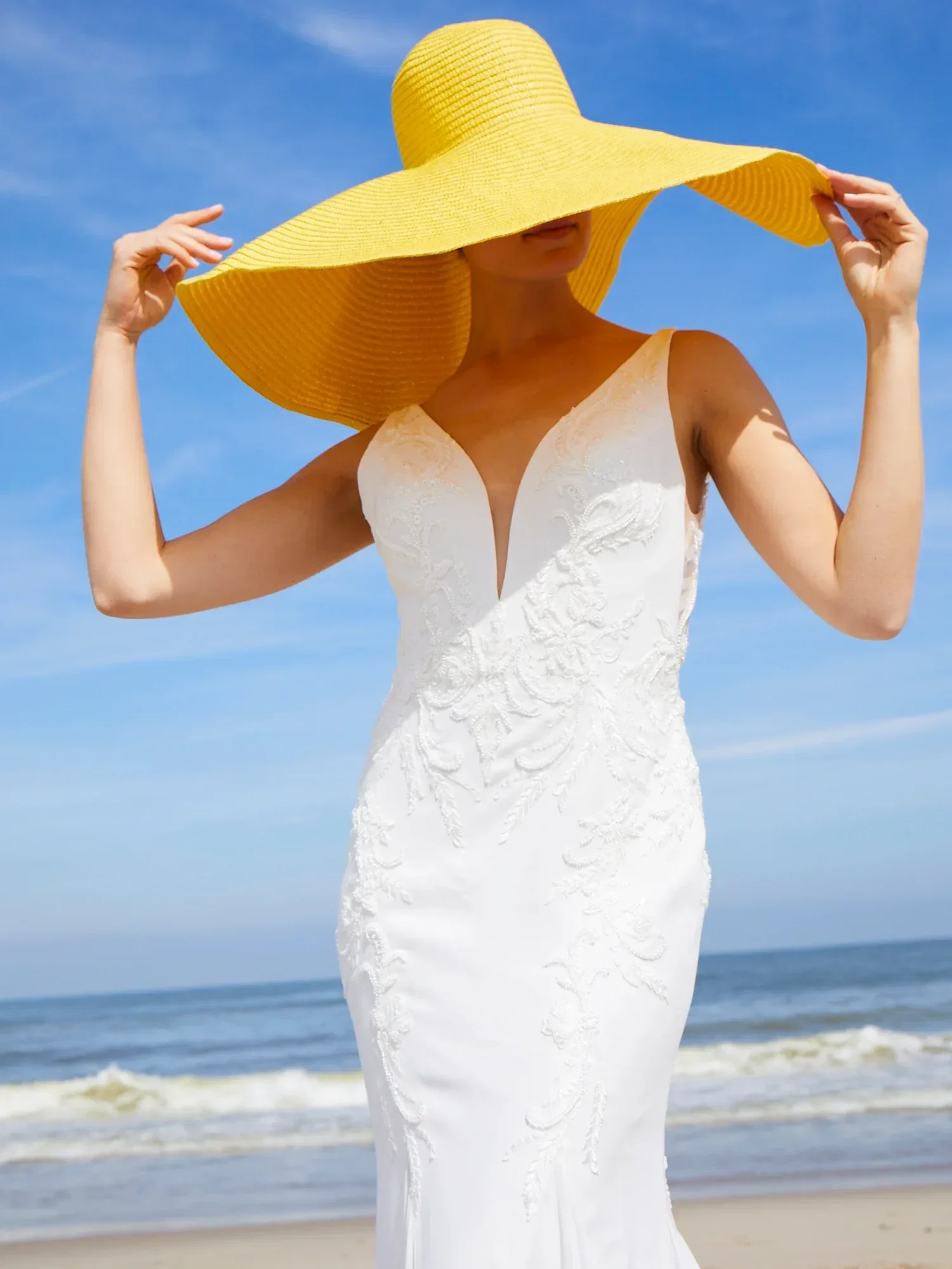 Eine Braut am Strand trägt ein weißes, elegant besticktes Kleid und einen auffälligen gelben Sonnenhut. Der Hut verdeckt teilweise ihr Gesicht.