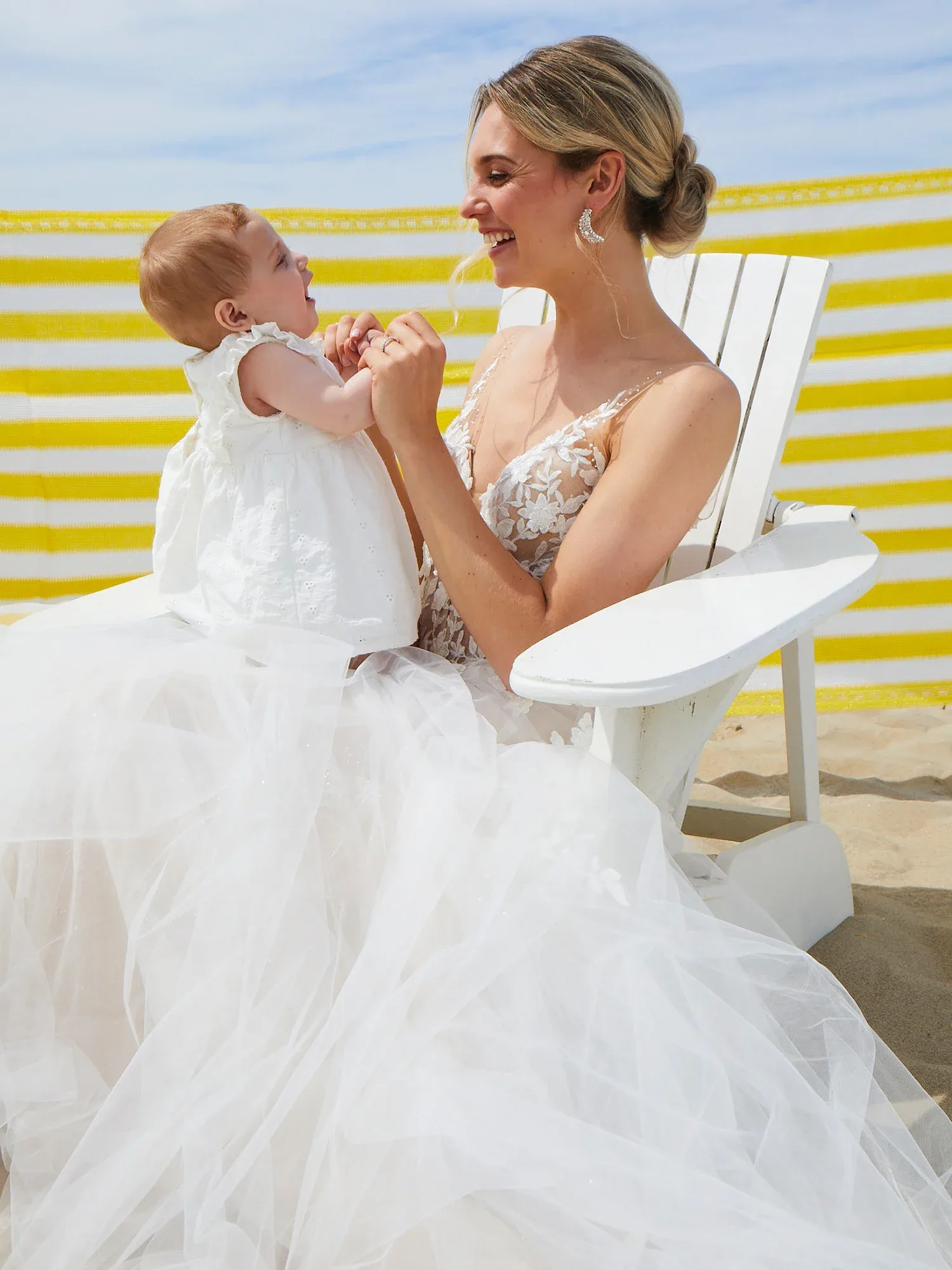 Eine Braut sitzt in einem weißen Sessel am Strand und hält ihr Baby auf dem Schoß. Beide lachen sich an, während die Braut ein voluminöses Spitzenkleid trägt.