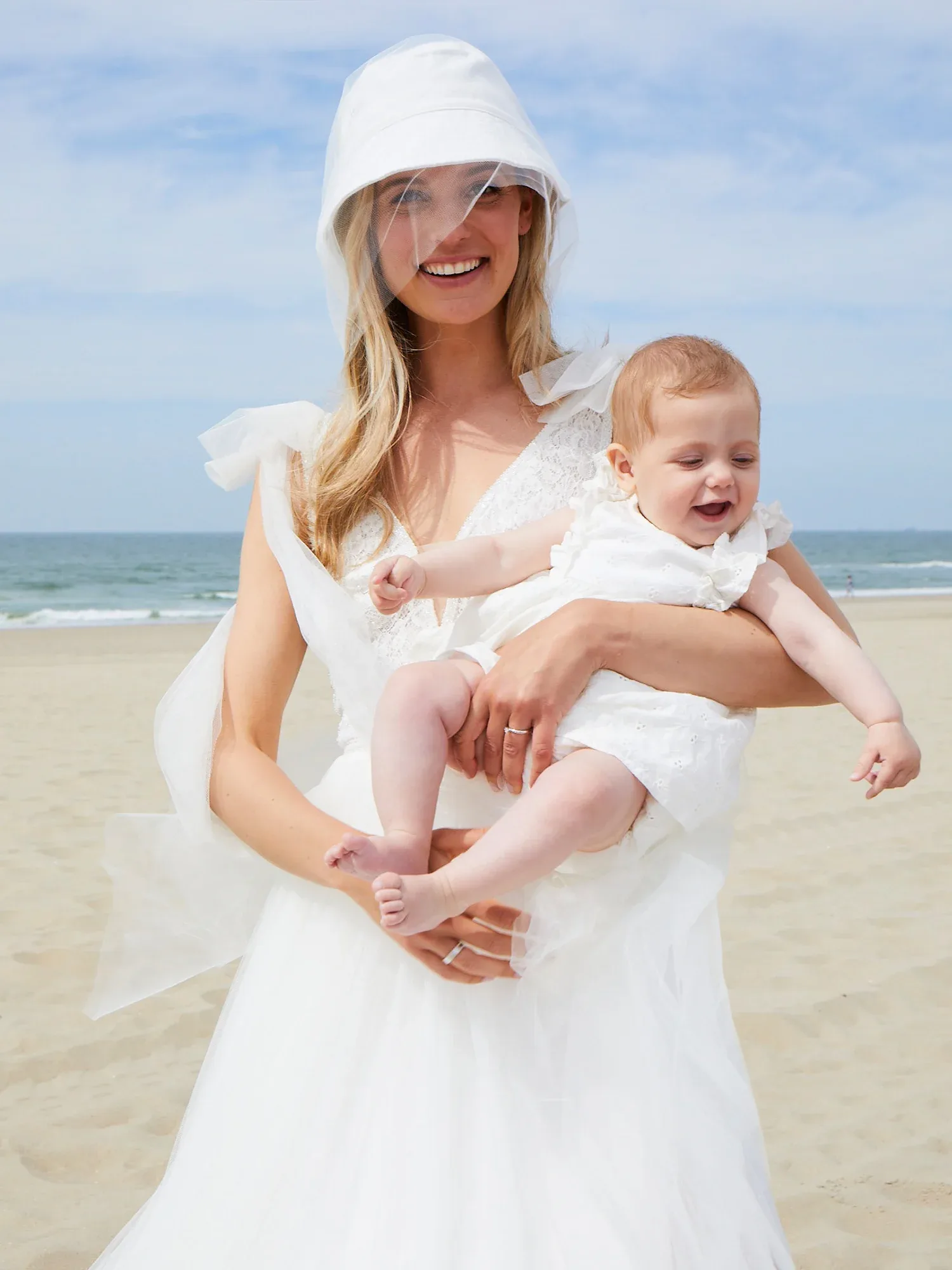 Eine Braut in einem weißen Kleid hält ein lachendes Baby auf dem Arm. Beide tragen passende weiße Outfits, die Szene spielt sich am Strand ab.