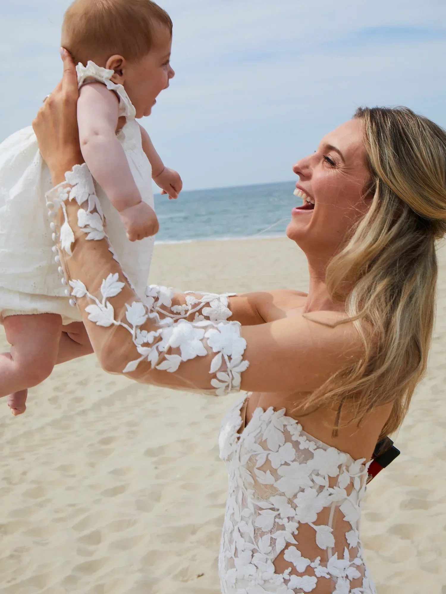 Eine Braut in einem weißen Spitzenkleid hebt lachend ihr Baby in die Luft, während sie am Strand steht. Beide wirken glücklich und entspannt.