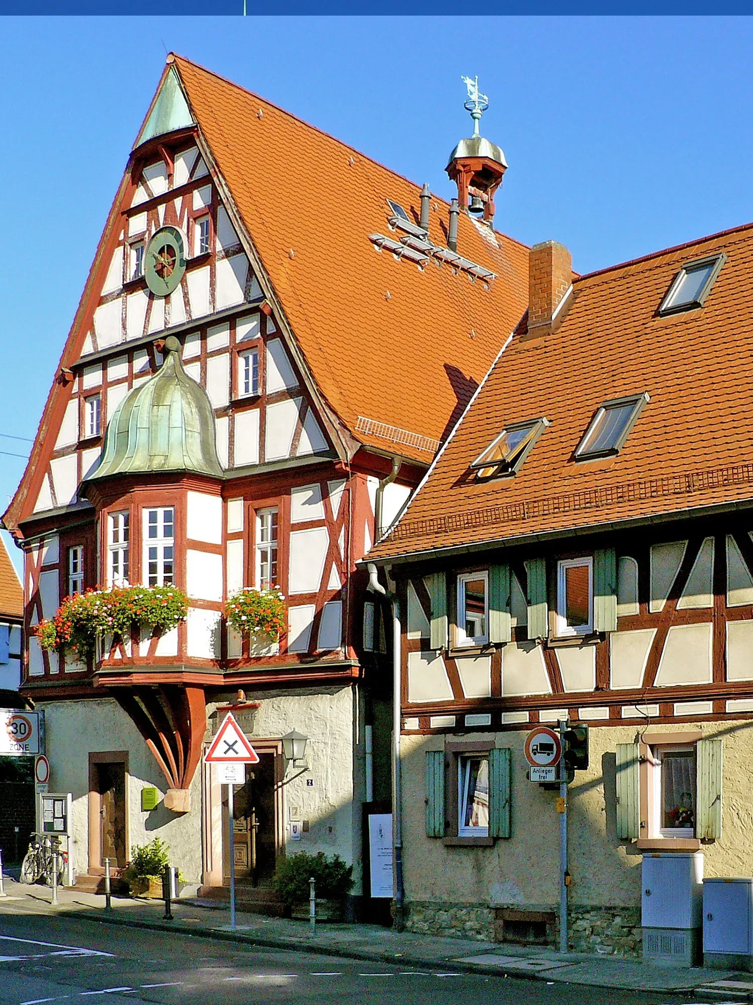Ein traditionelles Fachwerkhaus mit rotem Spitzdach, Seckbacher Rathaus genannt, strahlt im Sonnenlicht. Die Holzbalken und die weiße Fassade verleihen dem Gebäude einen historischen Charme. Daneben stehen kleinere Gebäude im ähnlichen Stil.
