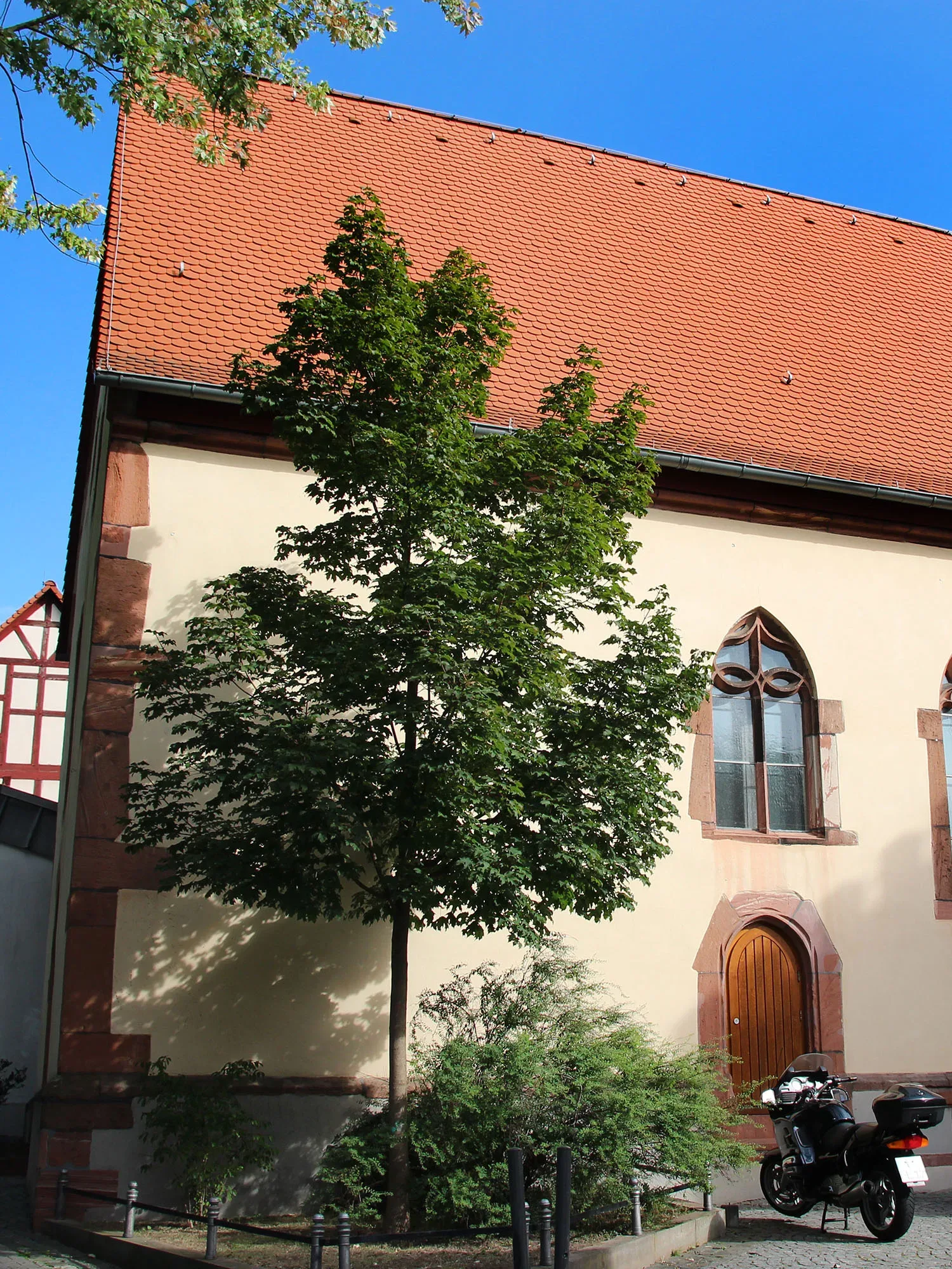 Die Nikolauskapelle mit schlichtem, cremefarbenem Mauerwerk und roten Ziegeln bildet eine ruhige Szenerie. Ein Baum steht neben dem Eingang, und im Vordergrund ist ein Motorrad geparkt.