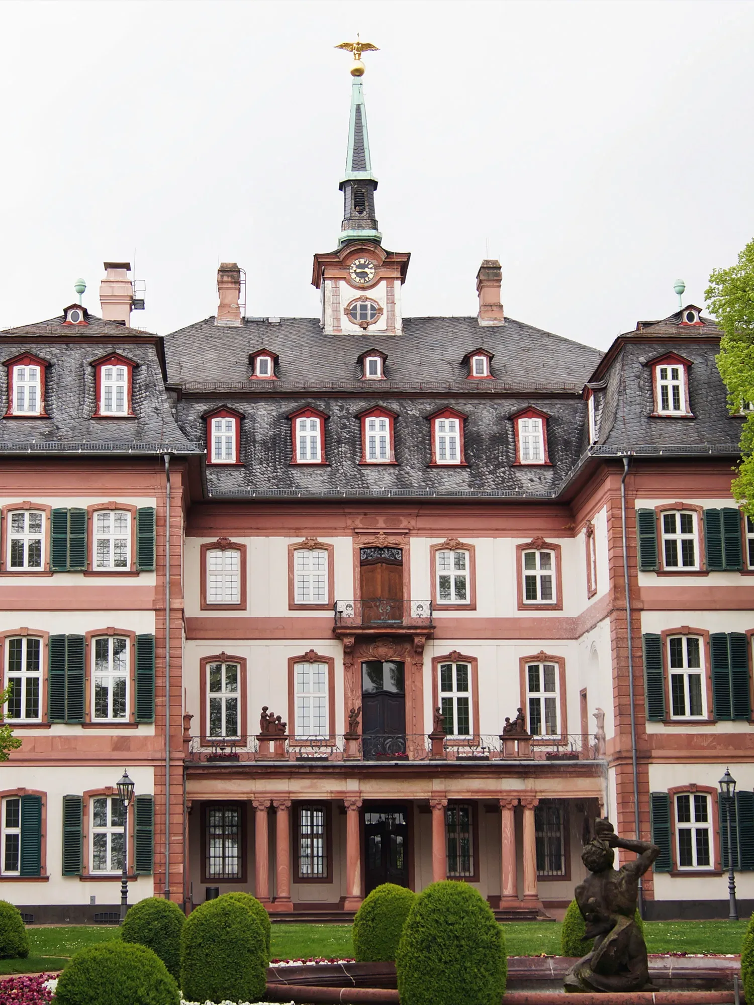 Der Bolongaropalast in Frankfurt-Höchst, ein imposantes Barockgebäude mit pastellfarbenen Fassaden, grünen Fensterläden und einem spitzen Turm mit Uhr und goldenem Adler. Im Vordergrund ist eine Skulptur im gepflegten Garten zu sehen.