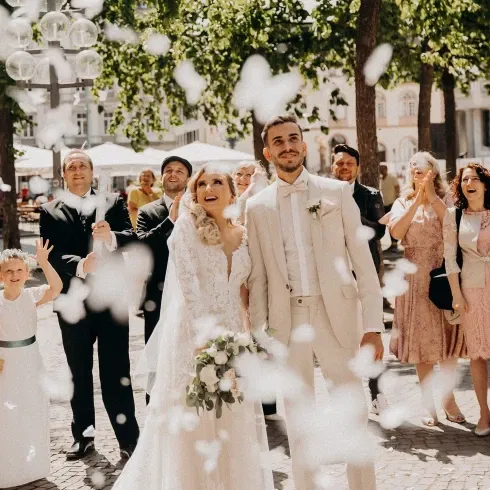 Ein frisch verheiratetes Paar steht vor einer Gruppe jubelnder Hochzeitsgäste, während weiße Blumenblätter in die Luft geworfen werden. Die Braut trägt ein elegantes Spitzenkleid und hält einen Blumenstrauß, während der Bräutigam in einem hellen Anzug lächelt. Kinder und Erwachsene feiern das Paar unter einem sonnigen Baum.