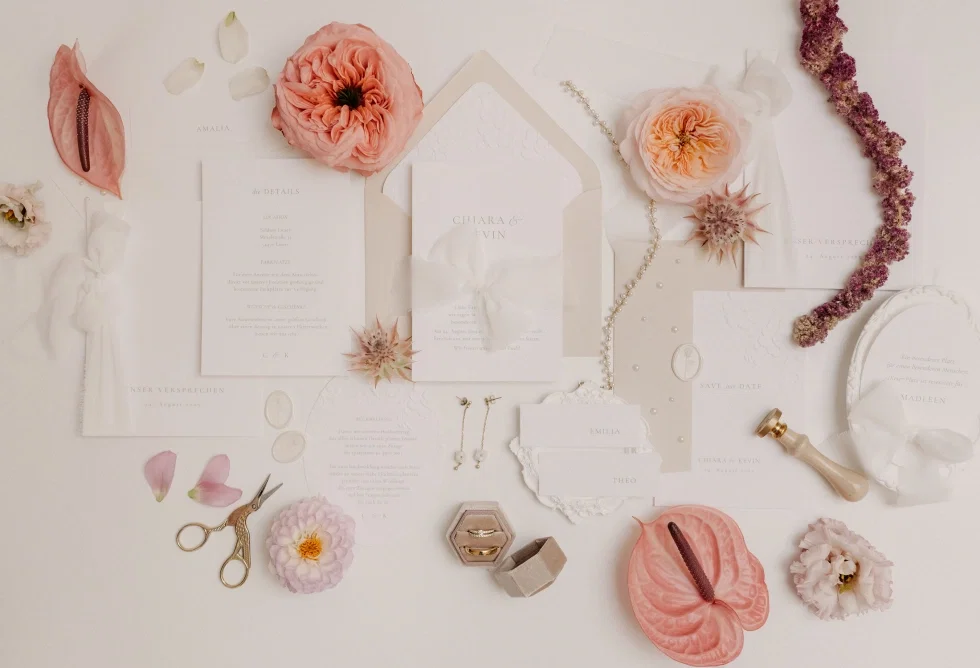 Romantisches Arrangement aus Hochzeitskarten, Umschlägen und floralen Elementen in Apricot- und Rosétönen, kombiniert mit Schere, Siegel und Schmuckdetails.