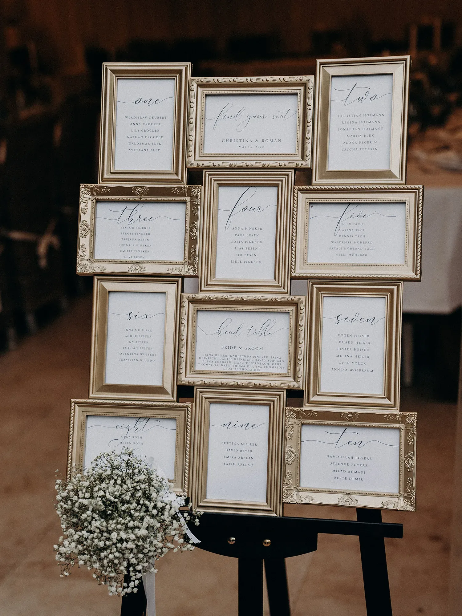 Ein eleganter Sitzplan für eine Hochzeit, präsentiert in goldenen Bilderrahmen. Die einzelnen Tischkarten sind in kalligrafischer Schrift gestaltet und zeigen die Namen der Gäste. Ein kleiner Blumenstrauß aus Schleierkraut schmückt den unteren Teil des Arrangements.
