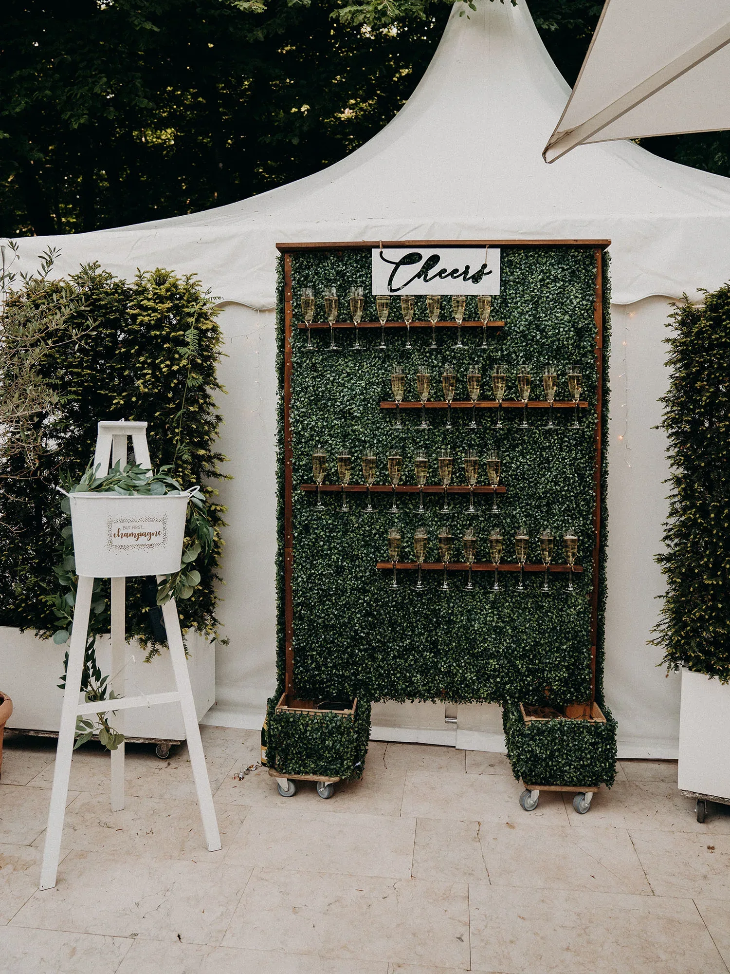 Eine stilvolle Champagner-Wand, dekoriert mit grünen Blättern und Gläsern, die auf Regalen präsentiert werden. Über der Wand hängt ein Schild mit der Aufschrift „Cheers“. Daneben steht ein weißer Eimerständer mit der Aufschrift „Champagne“, bereit für die Hochzeitsgäste.