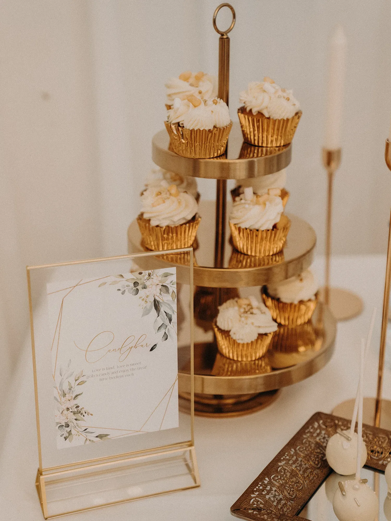 Ein eleganter Cupcake-Ständer mit goldenen Cupcakes und weißer Verzierung steht auf einem Sweet Table. Neben dem Ständer befindet sich ein dekoratives Schild mit floralen Elementen und der Aufschrift „Candy Bar“, das die Gäste zum Genießen der süßen Leckereien einlädt.