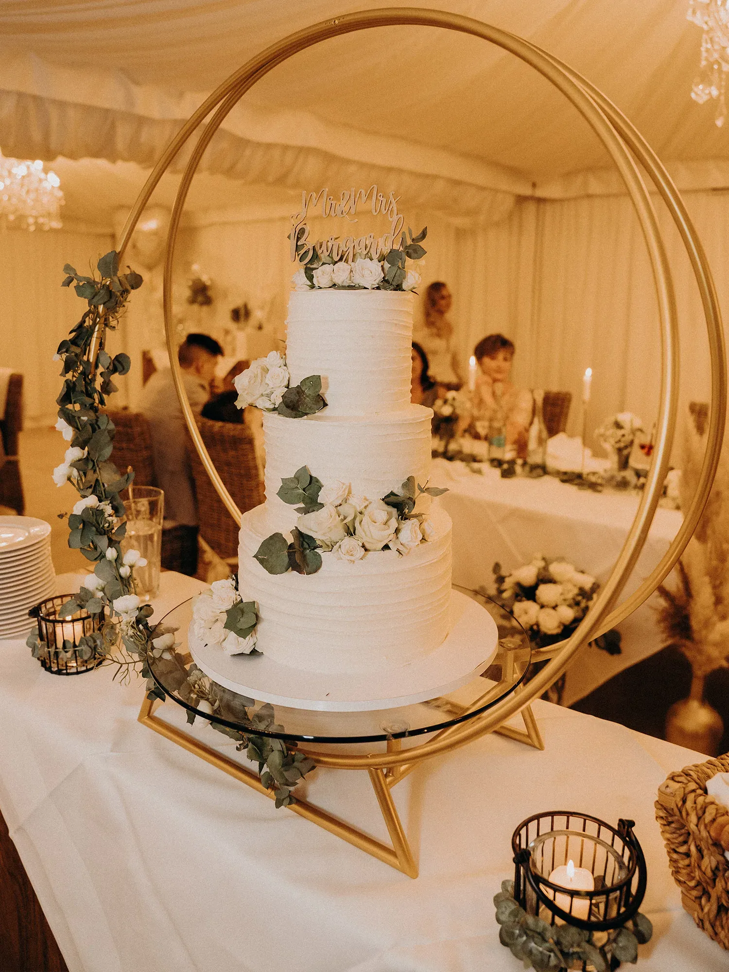 Eine elegante, dreistöckige Hochzeitstorte mit weißem Frosting, dekoriert mit weißen Rosen und Eukalyptusblättern. Die Torte steht in einem goldenen Ringständer und wird mit dem Schriftzug „Mr & Mrs“ auf der Spitze gekrönt. Um den Tisch herum sind Kerzen in dekorativen Haltern angeordnet, und im Hintergrund sind Hochzeitsgäste zu sehen.