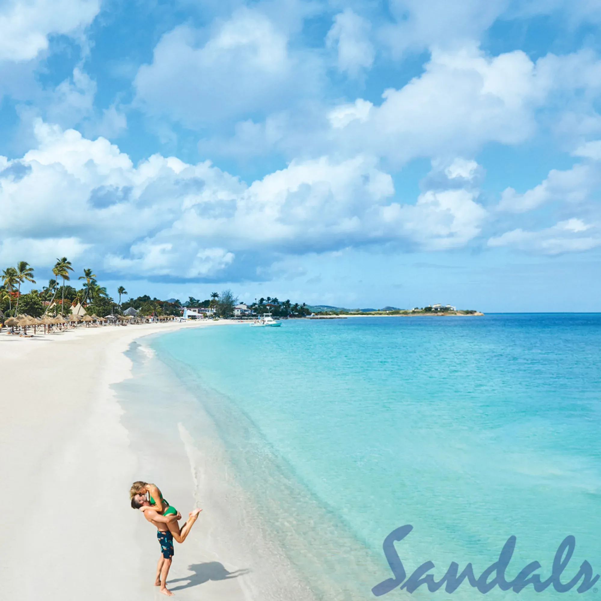 Ein Paar genießt einen romantischen Moment am weißen Sandstrand von Antigua. Der Mann hebt die Frau in die Luft, während sie am türkisblauen Wasser stehen. Der Himmel ist klar mit vereinzelten Wolken, und Palmen säumen den Strand in der Ferne.