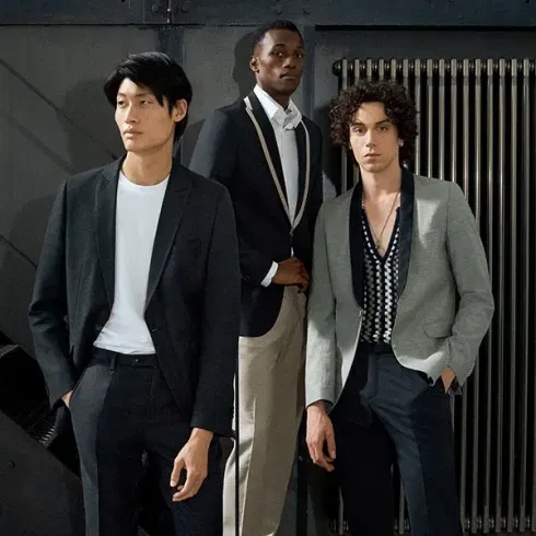 Drei Männer stehen in einem modernen, industriellen Raum. Sie tragen stilvolle Anzüge in Schwarz, Grau und Beige. Der Mann links trägt einen schwarzen Anzug mit einem weißen T-Shirt, während der Mann in der Mitte einen dunkelblauen Blazer und eine beigefarbene Hose trägt. Der dritte Mann rechts trägt einen grauen Blazer mit einem gemusterten Hemd darunter.