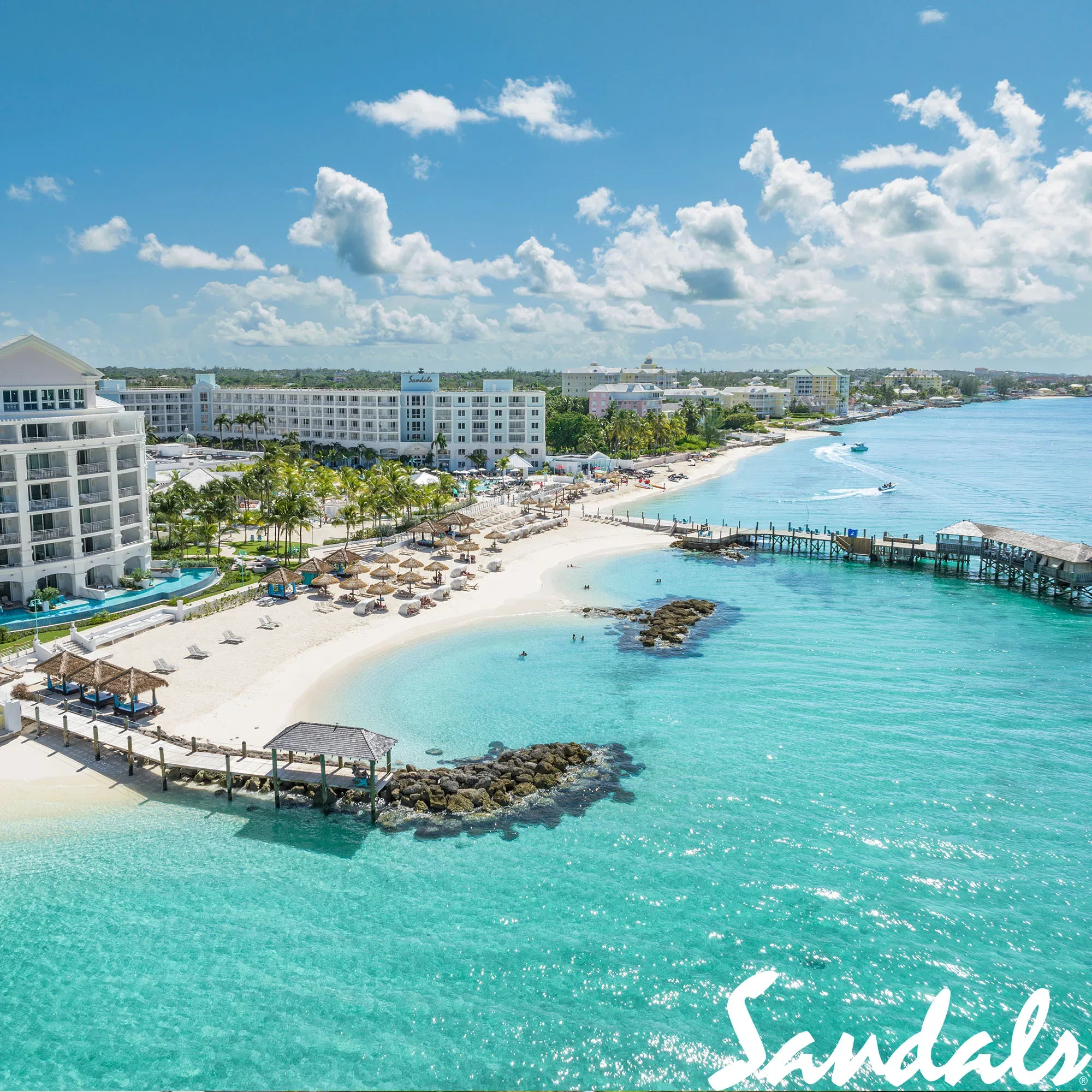 Eine Vogelperspektive auf den weißen Sandstrand und das türkisfarbene Wasser eines Resorts auf den Bahamas. Das Resort ist von Palmen und Sonnenschirmen gesäumt, und das klare Meer lädt zum Schwimmen und Entspannen ein.