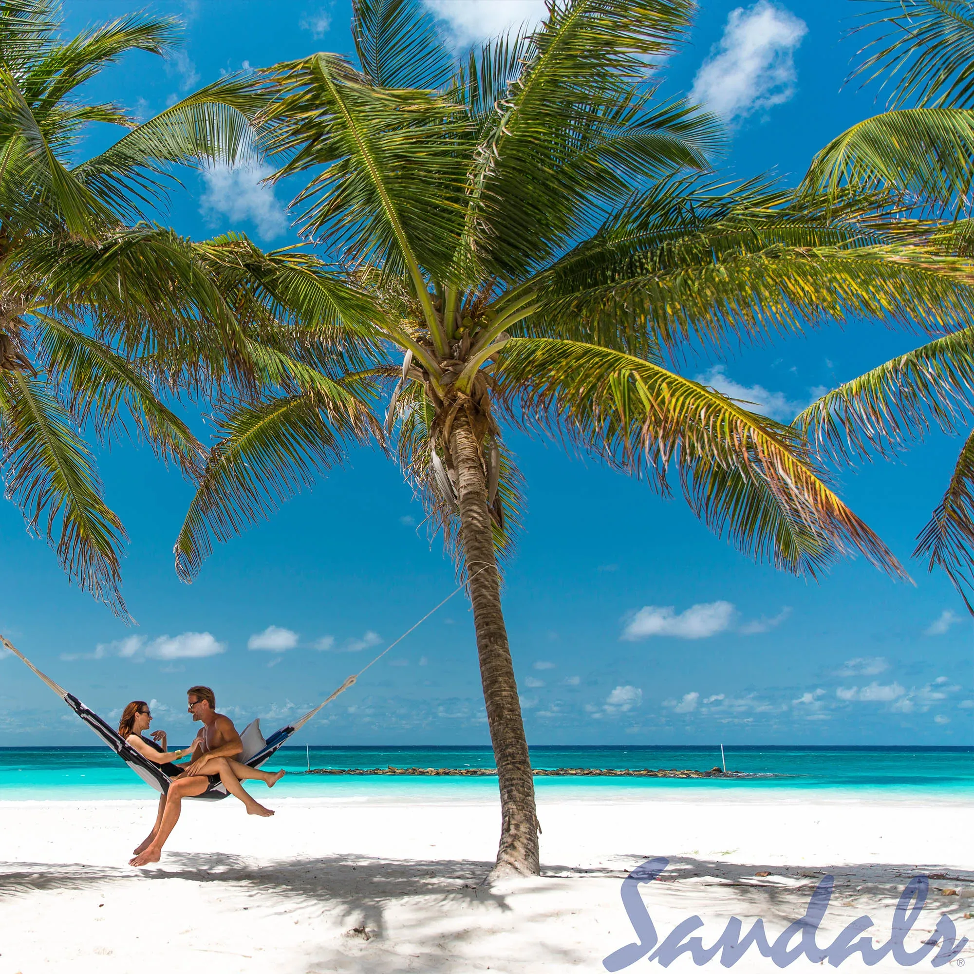 Ein Paar entspannt in einer Hängematte unter Palmen am weißen Sandstrand von Barbados. Der klare blaue Himmel und das türkisfarbene Meer schaffen eine idyllische, tropische Kulisse.
