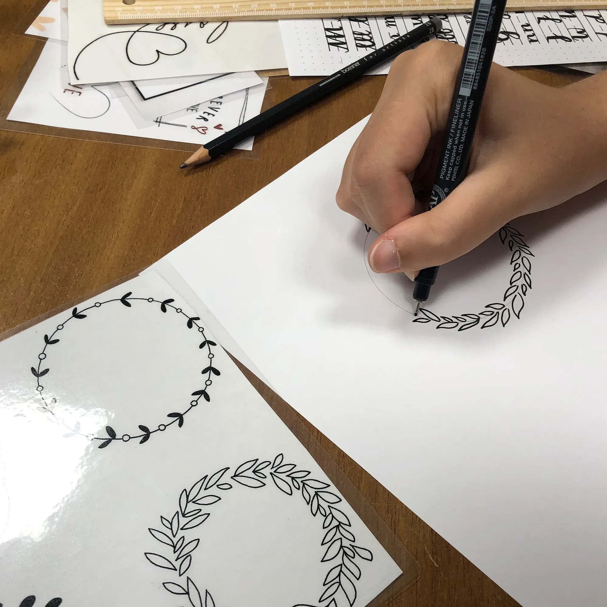 Nahaufnahme einer Hand, die mit einem Pinselstift einen Blattkranz auf Papier skizziert. Auf dem Tisch liegen andere Muster und Vorlagen für kreative Handlettering-Designs.