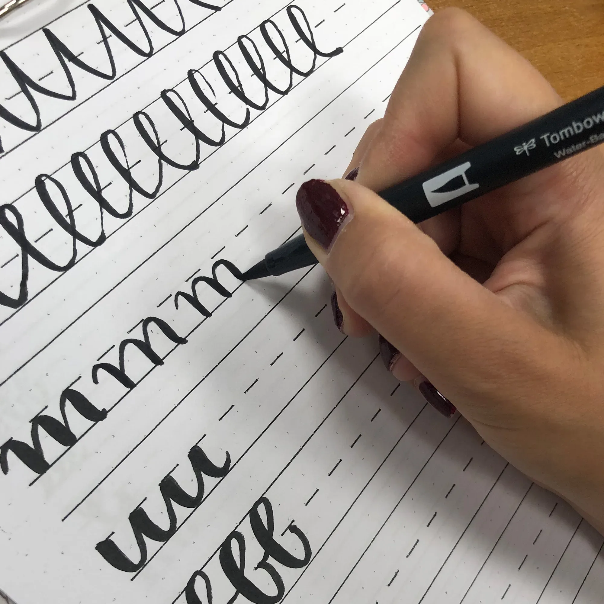 Eine Hand mit dunkelrot lackierten Fingernägeln übt Handlettering auf einem Arbeitsblatt, indem sie Buchstaben mit einem schwarzen Pinselstift nachzieht. Die Übungszeilen zeigen elegante, geschwungene Buchstabenformen.