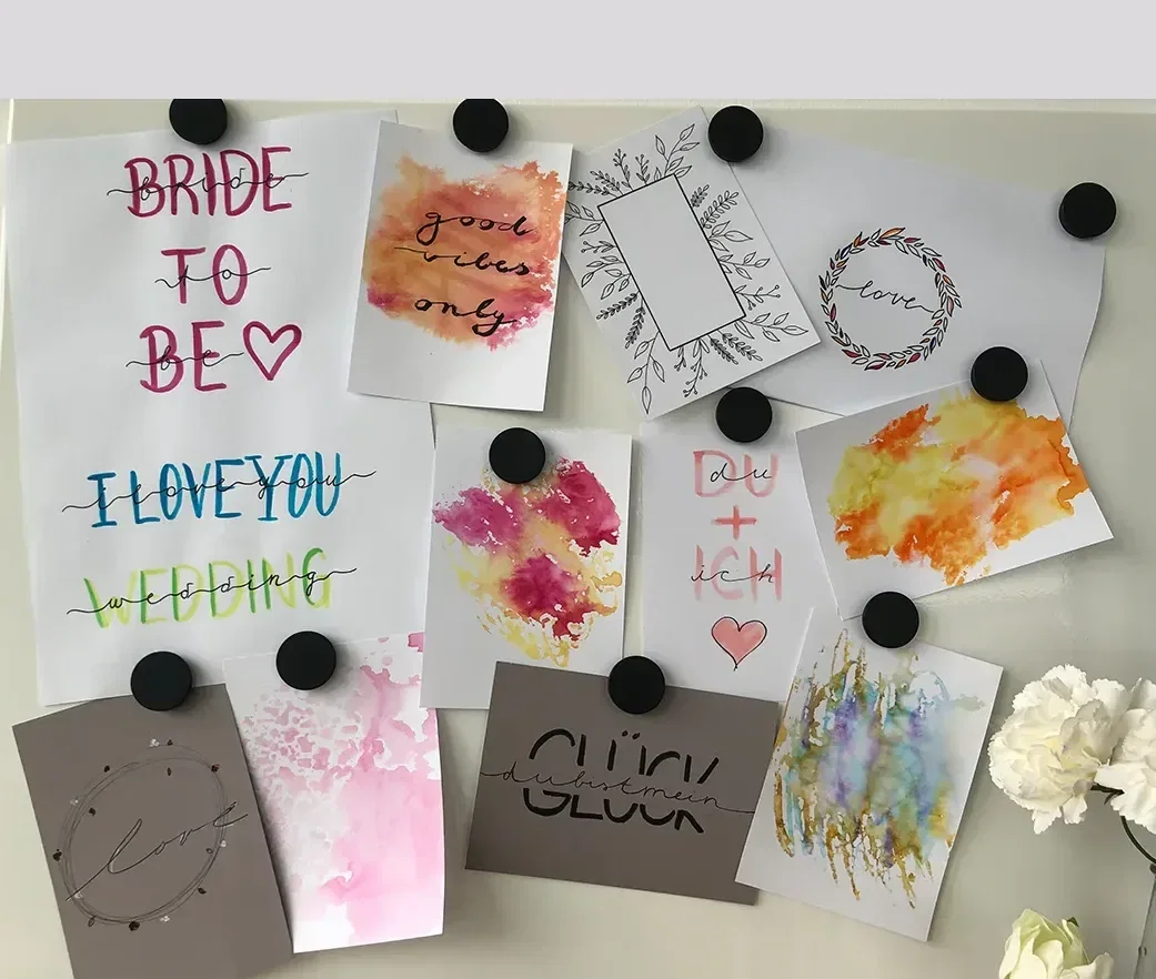 Eine Pinnwand mit verschiedenen kreativen Handlettering- und Aquarell-Arbeiten. Schilder mit Schriftzügen wie "Bride to Be" und "I Love You" sind zu sehen.