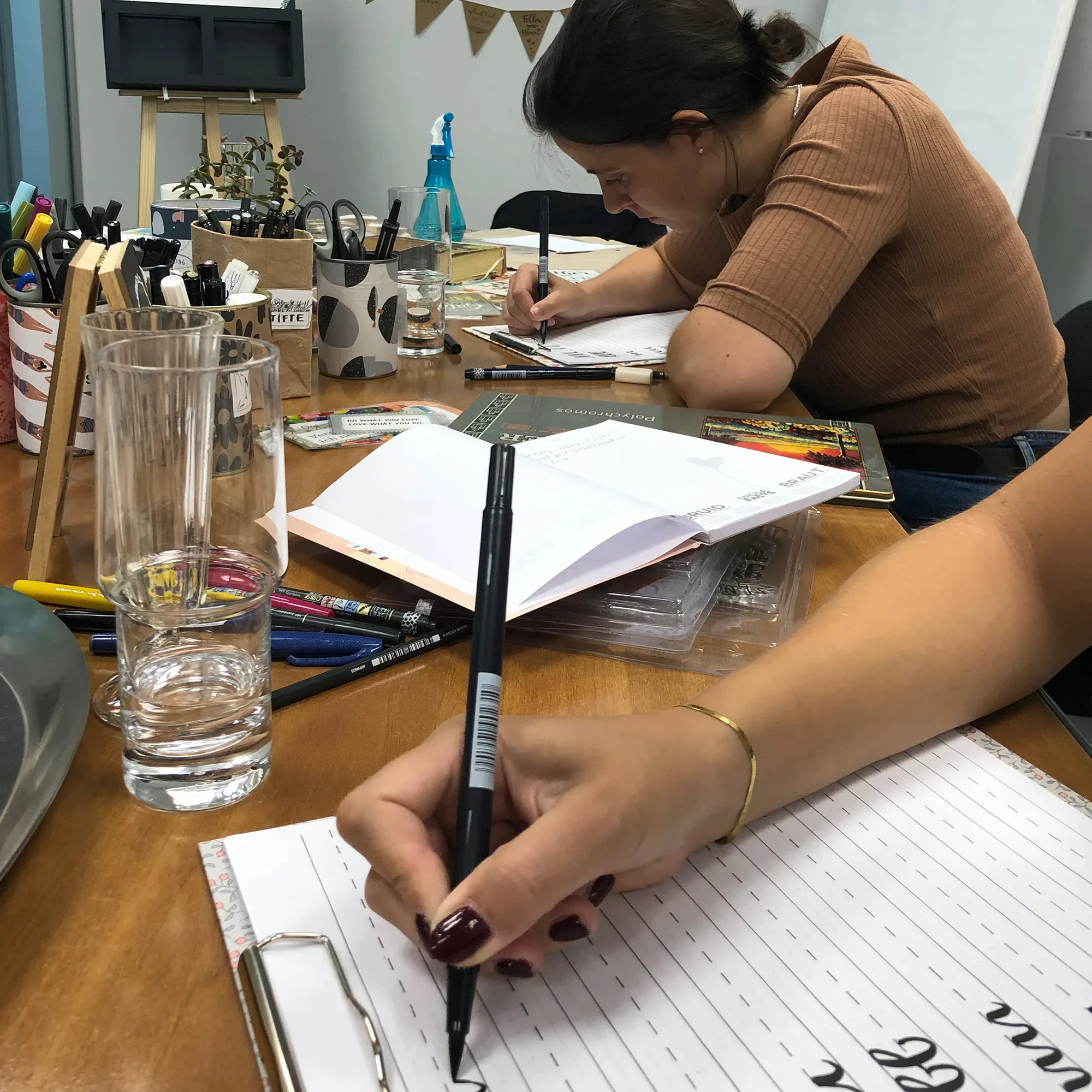 Eine Gruppe von Teilnehmern sitzt an einem Holztisch und übt Handlettering. Auf dem Tisch liegen Stifte, Pinsel, Gläser und Handlettering-Arbeitsblätter. Im Hintergrund arbeitet eine Frau konzentriert an ihren Buchstaben.