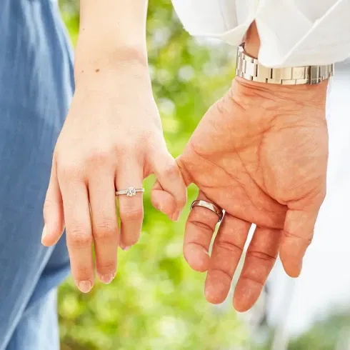 Nahaufnahme zweier Hände, die sich leicht berühren. Die linke Hand trägt einen funkelnden Verlobungsring mit einem Diamanten, während die rechte Hand einen schlichten Ehering trägt. Im Hintergrund ist verschwommenes Grün zu sehen, was eine ruhige, natürliche Atmosphäre vermittelt.