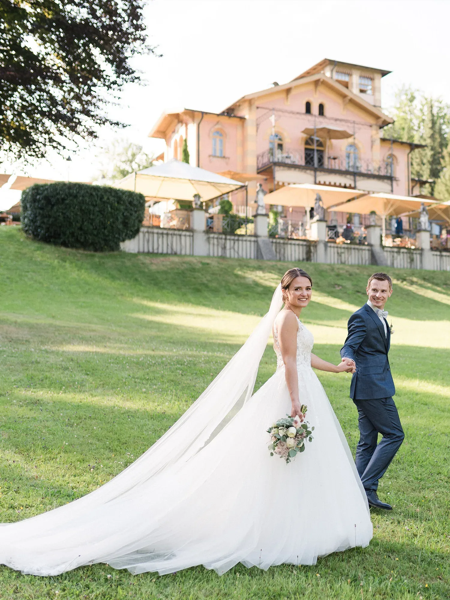Ein glückliches Brautpaar spaziert Hand in Hand über eine grüne Wiese vor einer charmanten Villa. Die Braut trägt ein weißes Kleid mit langem Schleier und hält einen Blumenstrauß, während der Bräutigam einen dunkelblauen Anzug trägt.