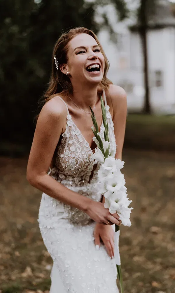 Lachende Braut in einem perlenbesetzten Hochzeitskleid, die eine Gladiolenblume in der Hand hält und im Freien steht.