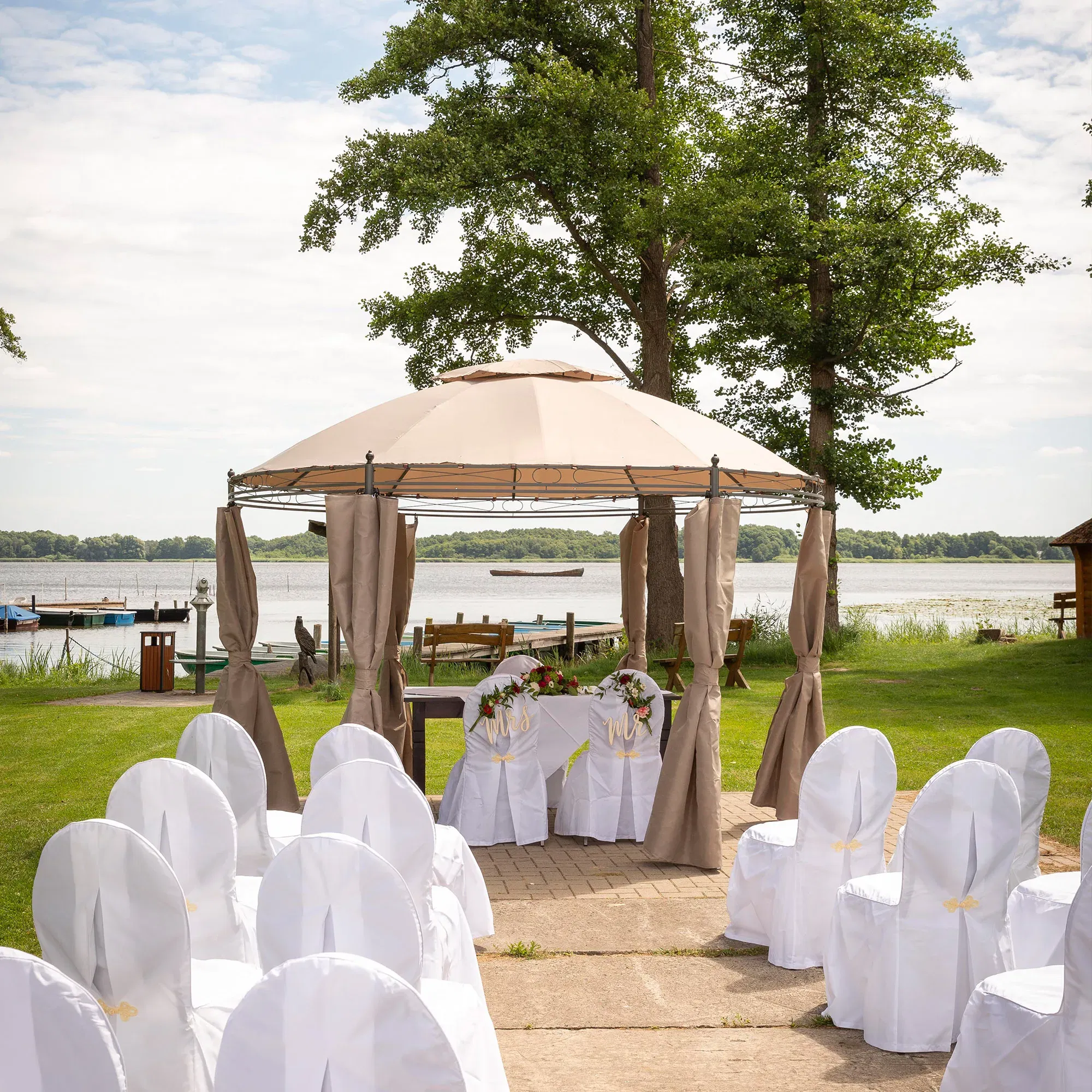 Eine romantische Hochzeitszeremonie unter einem Pavillon am Seeufer, umgeben von Bäumen und Stühlen, die mit weißen Hussen bedeckt sind. Der Blick auf den ruhigen See im Hintergrund verleiht der Szene eine idyllische Atmosphäre.