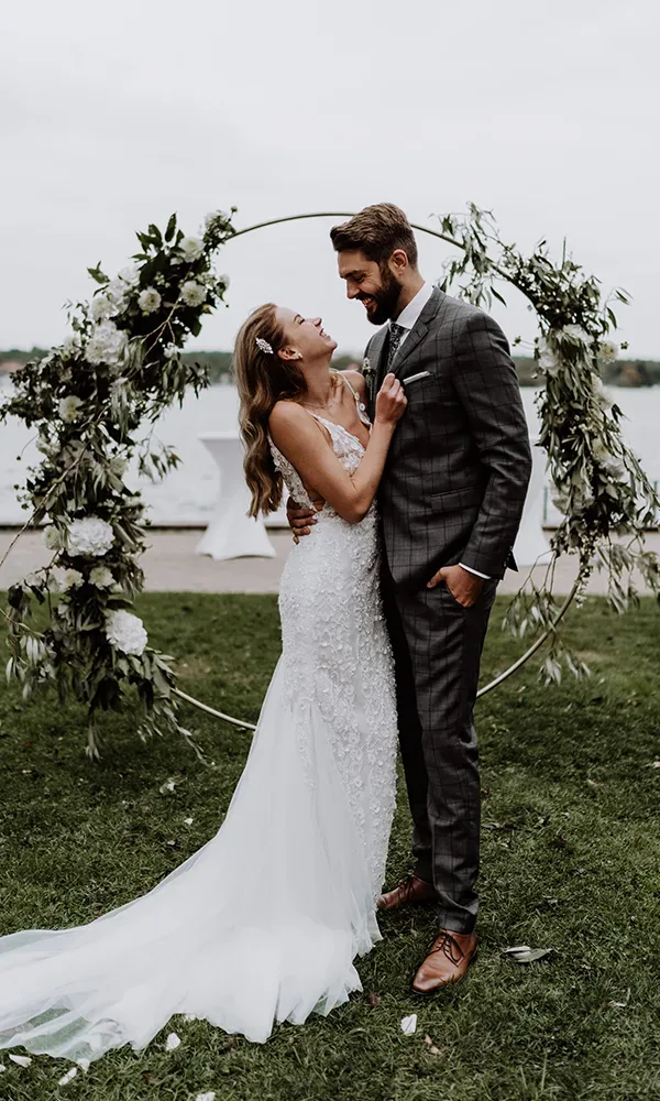 Hochzeitspaar in eleganter Kleidung vor einem Blumenbogen am Wasser. Die Braut in einem perlenbesetzten Kleid, der Bräutigam im grauen Anzug.