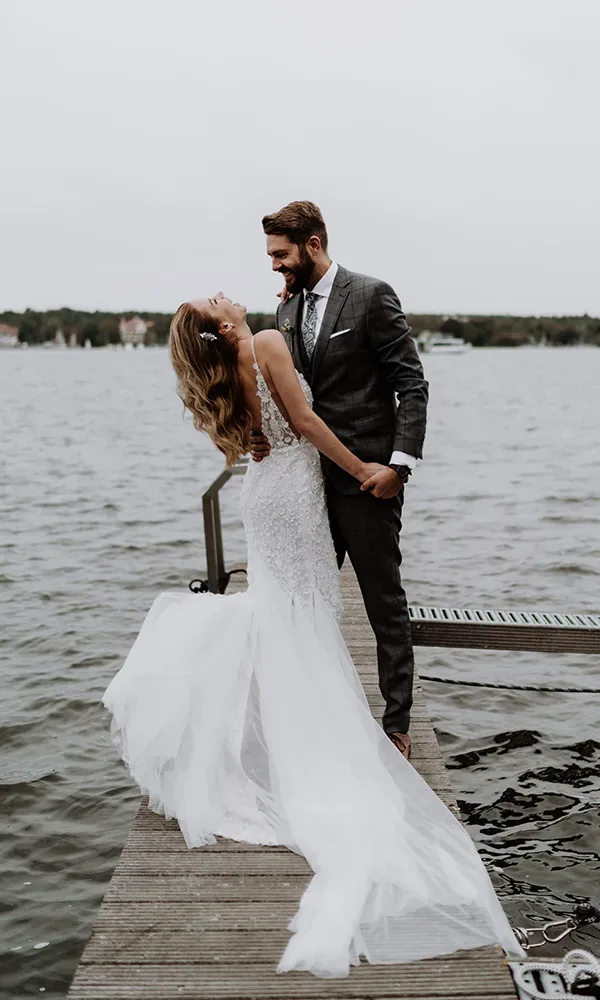 Ein glückliches Hochzeitspaar, das verliebt auf einem Steg am See steht und in die Augen sieht.