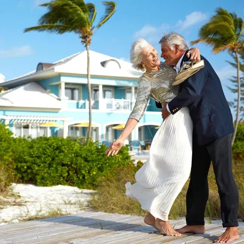 Barfuß und glücklich tanzt ein älteres Ehepaar auf einem Holzweg, mit Wind in den Haaren und einer tropischen Villa im Hintergrund.