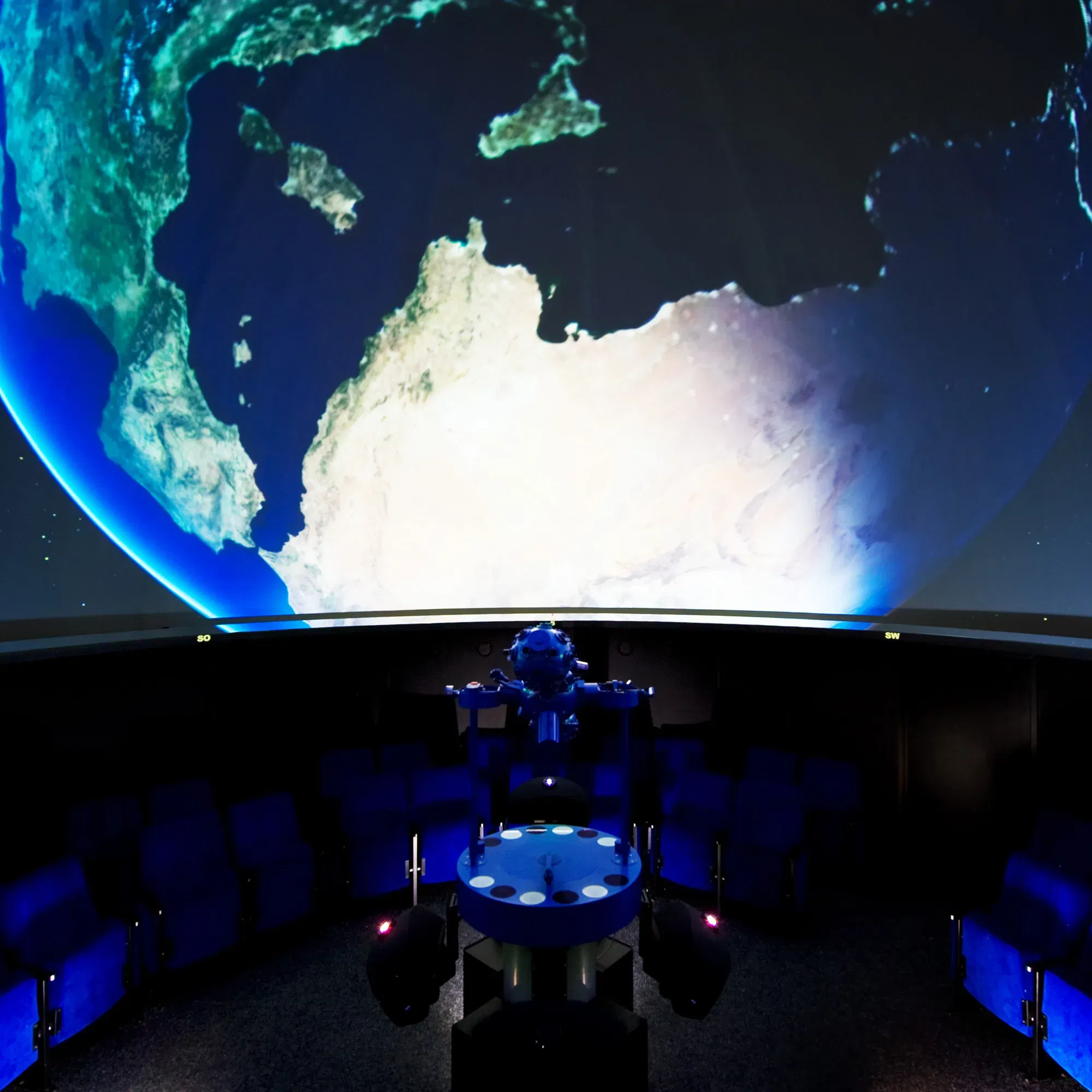 Blick in ein Planetarium mit einem hochmodernen Projektor, der das Bild der Erde auf die große Kuppeldecke projiziert. Um den Projektor herum befinden sich Sitzreihen für die Zuschauer.