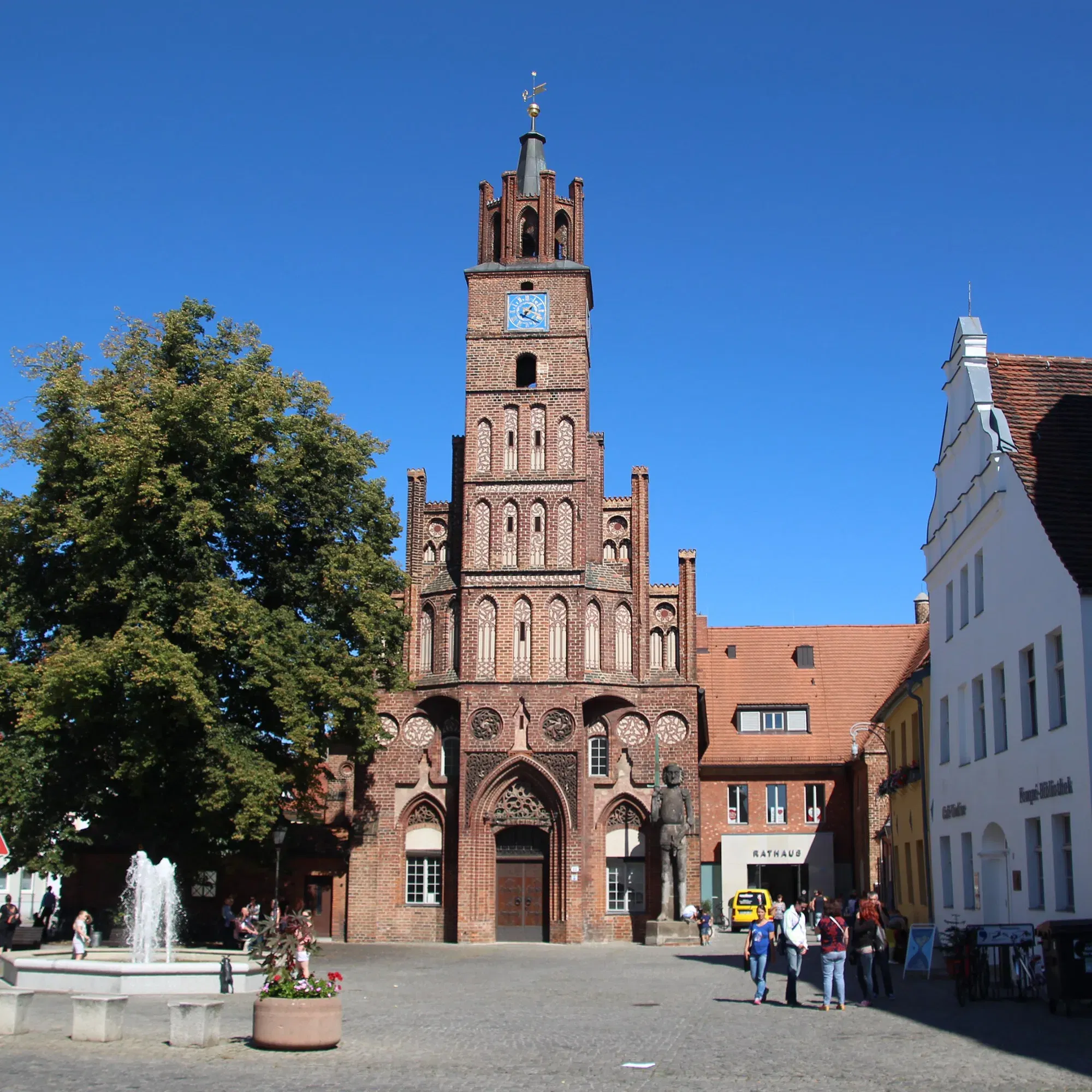 Das historische Rathaus von Brandenburg mit seiner markanten Backsteinfassade und dem hohen Turm steht im Zentrum des Platzes, flankiert von einem großen Baum und umliegenden Gebäuden. Vor dem Rathaus befindet sich ein kleiner Brunnen.