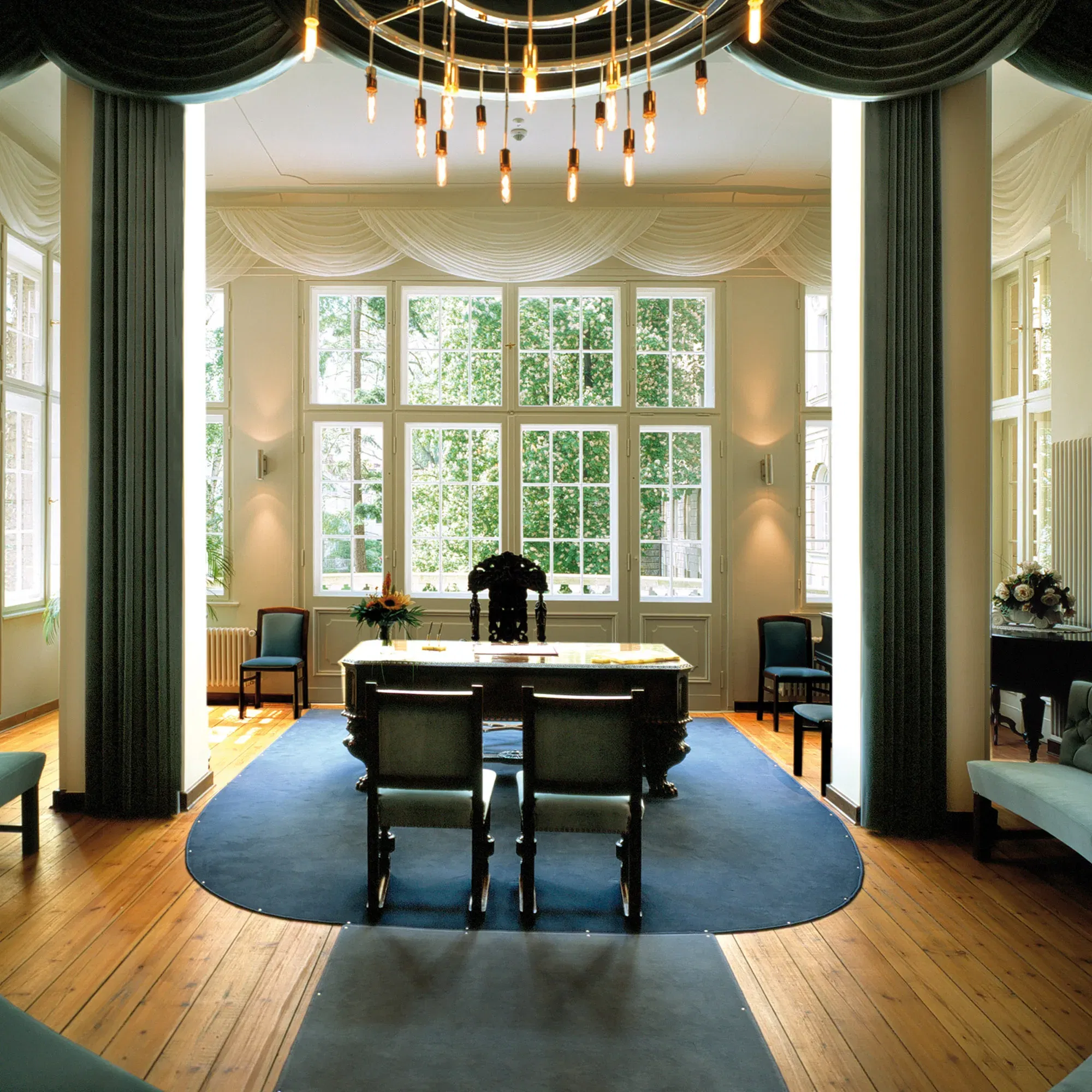 Ein großzügiger Trausaal mit hohen Fenstern, durch die grüne Bäume zu sehen sind. In der Mitte steht ein großer Tisch, flankiert von Stühlen, und der Raum wird von einem modernen, kreisförmigen Kronleuchter beleuchtet.