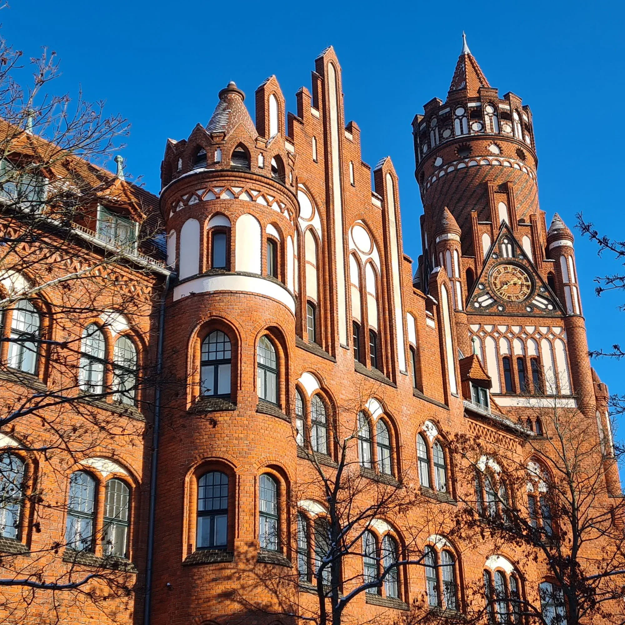 Das Rathaus Schmargendorf beeindruckt mit seiner roten Backsteinfassade und den architektonisch markanten Türmen und Bögen. Der klare blaue Himmel lässt die gotischen Elemente des Gebäudes besonders hervortreten.