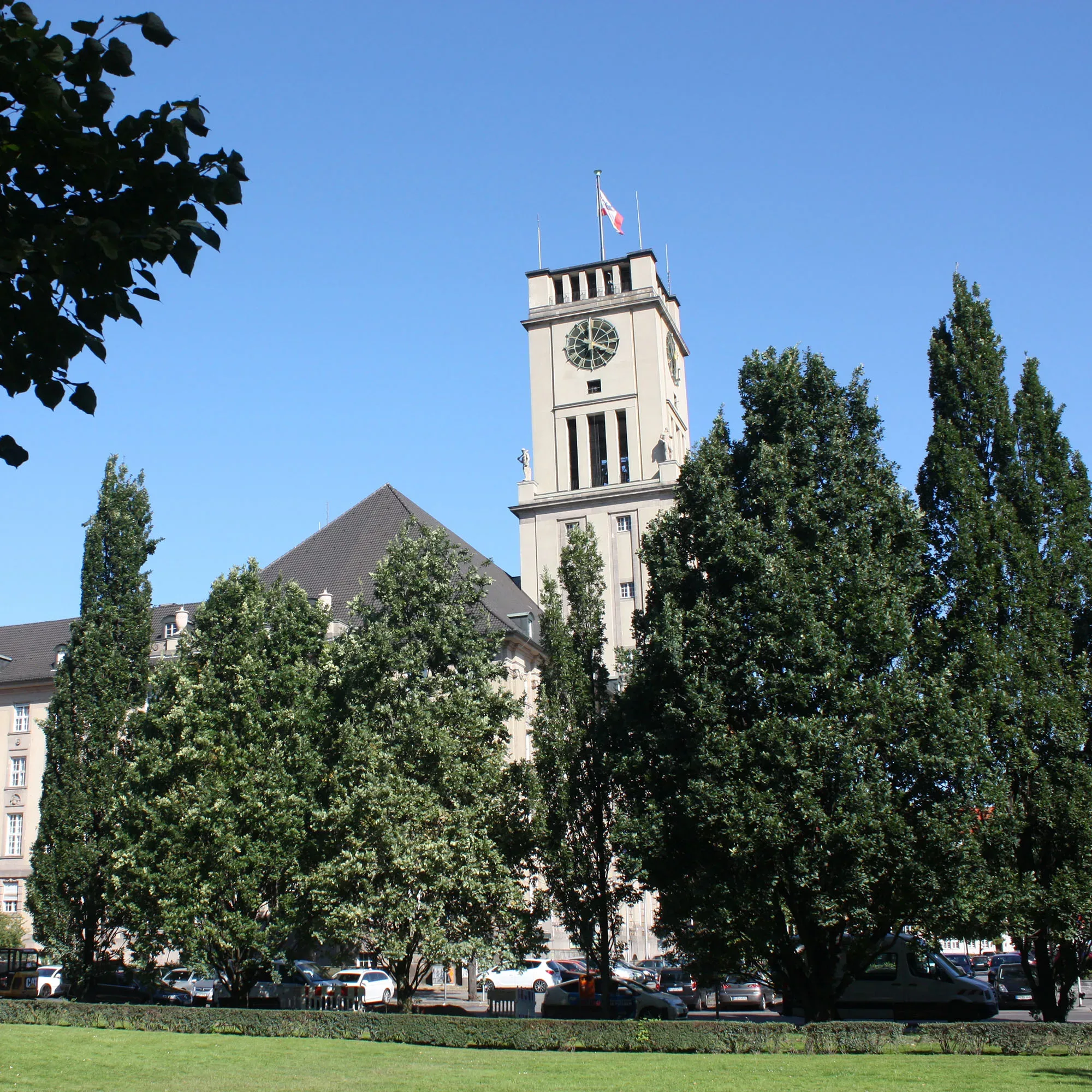 Das historische Rathaus Schöneberg, bekannt für seinen hohen Turm mit der Uhr, erhebt sich über die Baumkronen eines angrenzenden Parks. Das markante Gebäude ist von grünen Bäumen umgeben und strahlt an einem klaren, blauen Himmel.