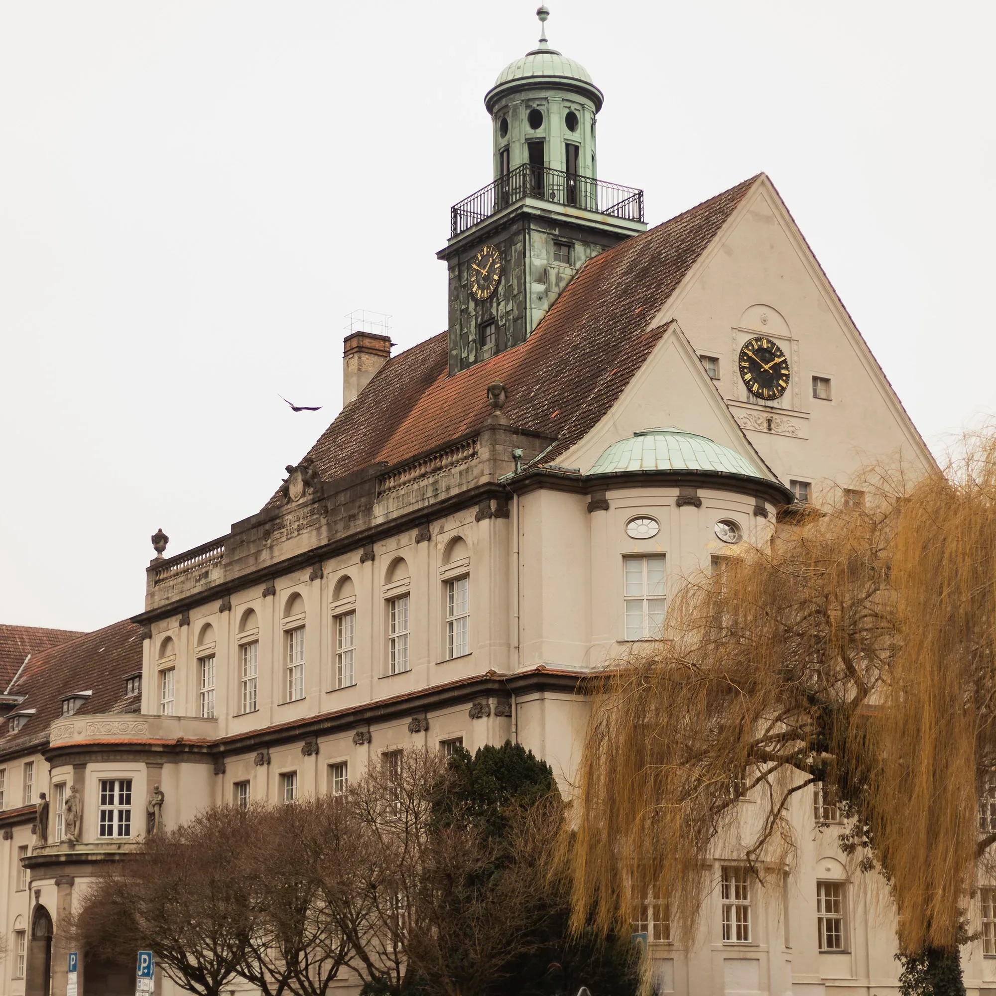 Das Rathaus Treptow, ein imposantes historisches Gebäude mit einer großen Turmuhr und Kupferdach, thront majestätisch über den Bäumen. Der rote Ziegeldach und die klassischen Fassadenelemente spiegeln die historische Bedeutung des Gebäudes wider.