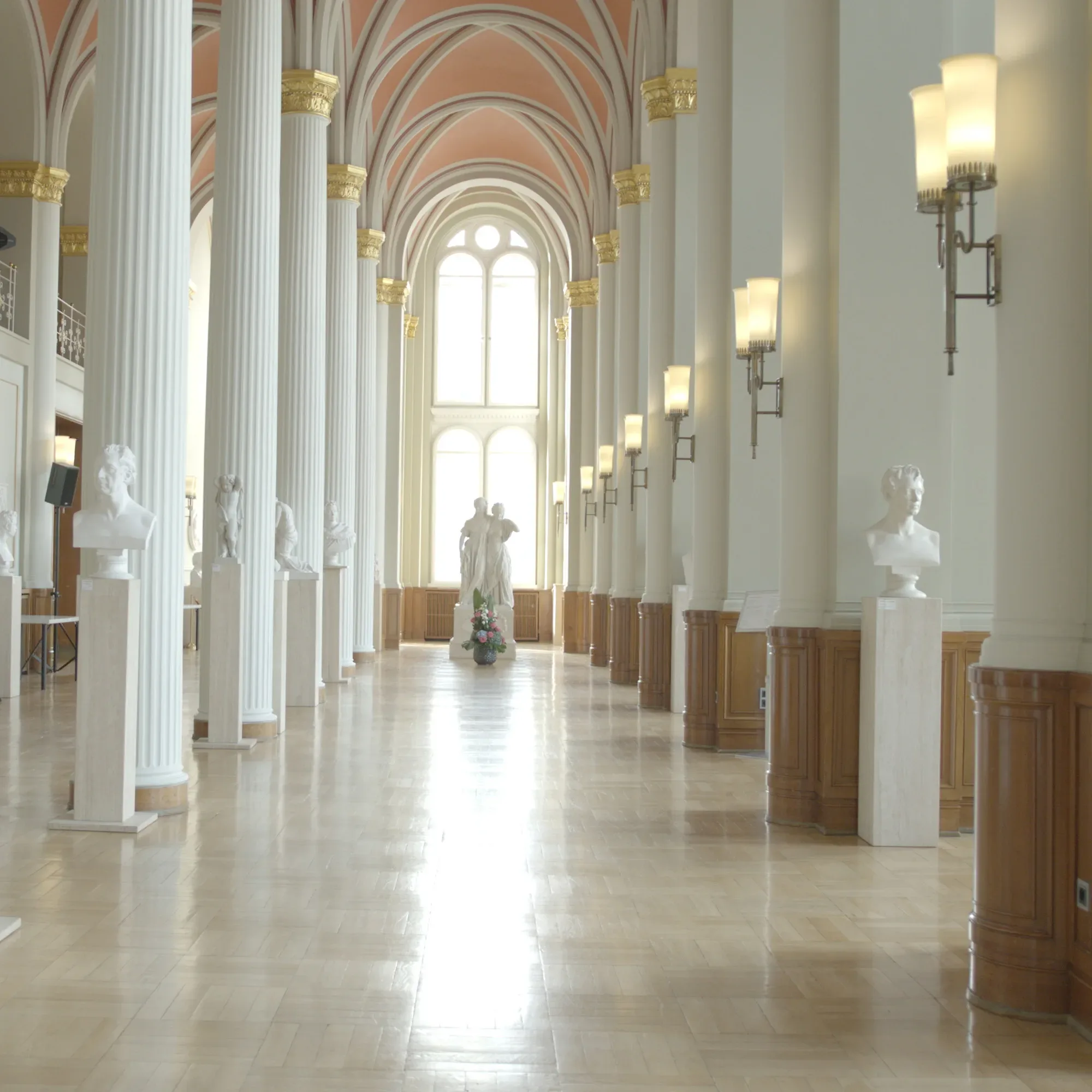 Die prächtige Innenansicht des Roten Rathauses mit hohen Säulen und Marmorbüsten, die entlang des Flurs aufgestellt sind. Im Hintergrund strömt Licht durch die großen Bogenfenster, während die dekorativen Deckenbögen in zarten Rosatönen den Raum schmücken.