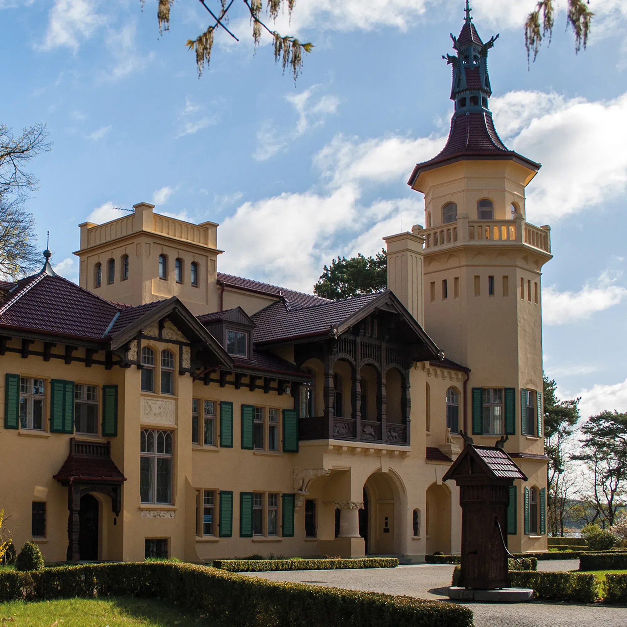 Das Schloss Hubertushöhe, ein historisches Gebäude mit einem markanten Turm und grünen Fensterläden, thront majestätisch unter einem blauen Himmel mit einigen Wolken. Die gepflegten Gärten umrahmen das Schloss stilvoll.