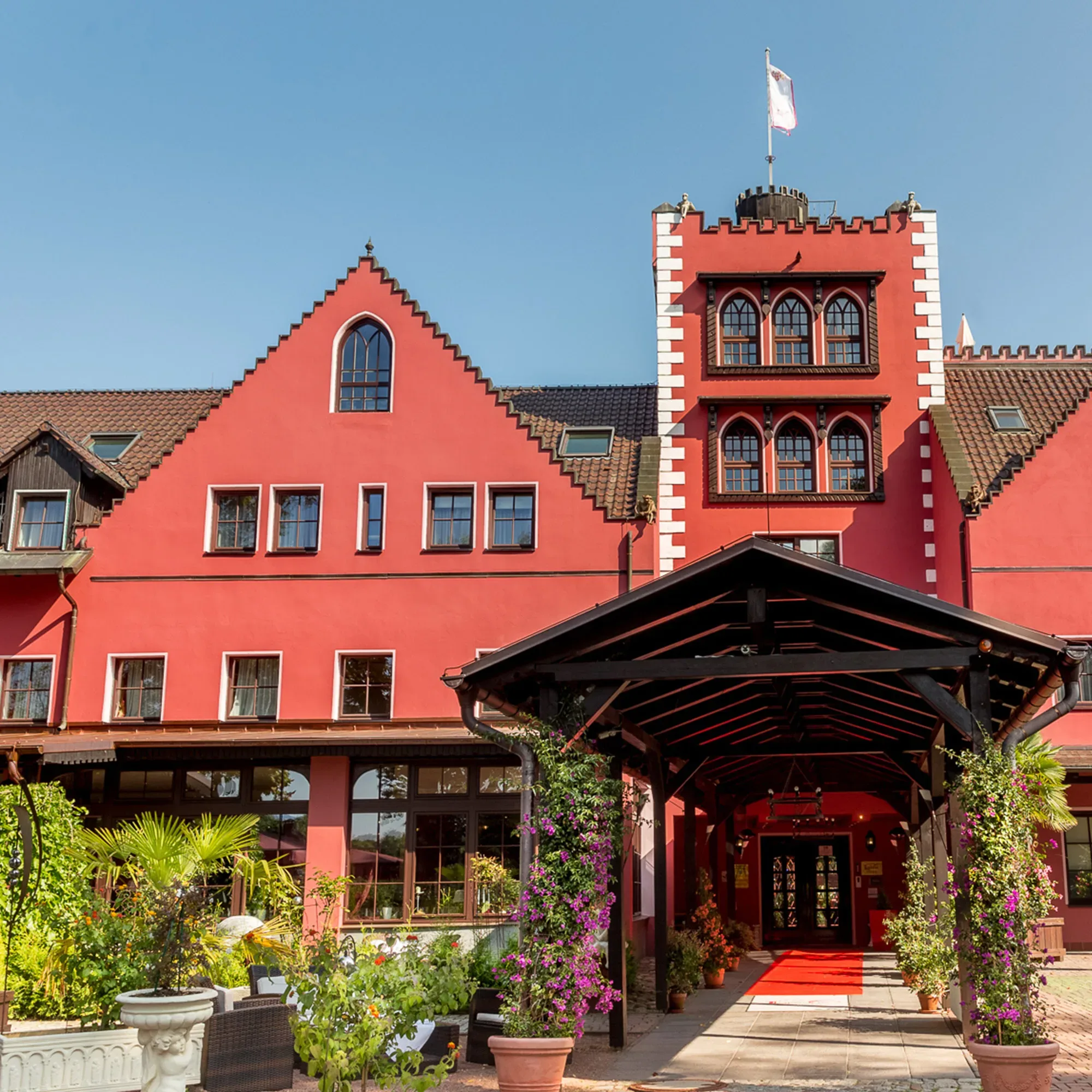 Das Lakeside Hotel, ein markantes Gebäude mit roter Fassade und einem überdachten Eingang, ist umgeben von blühenden Pflanzen. Der rote Teppich führt unter einem blumengeschmückten Baldachin hindurch.