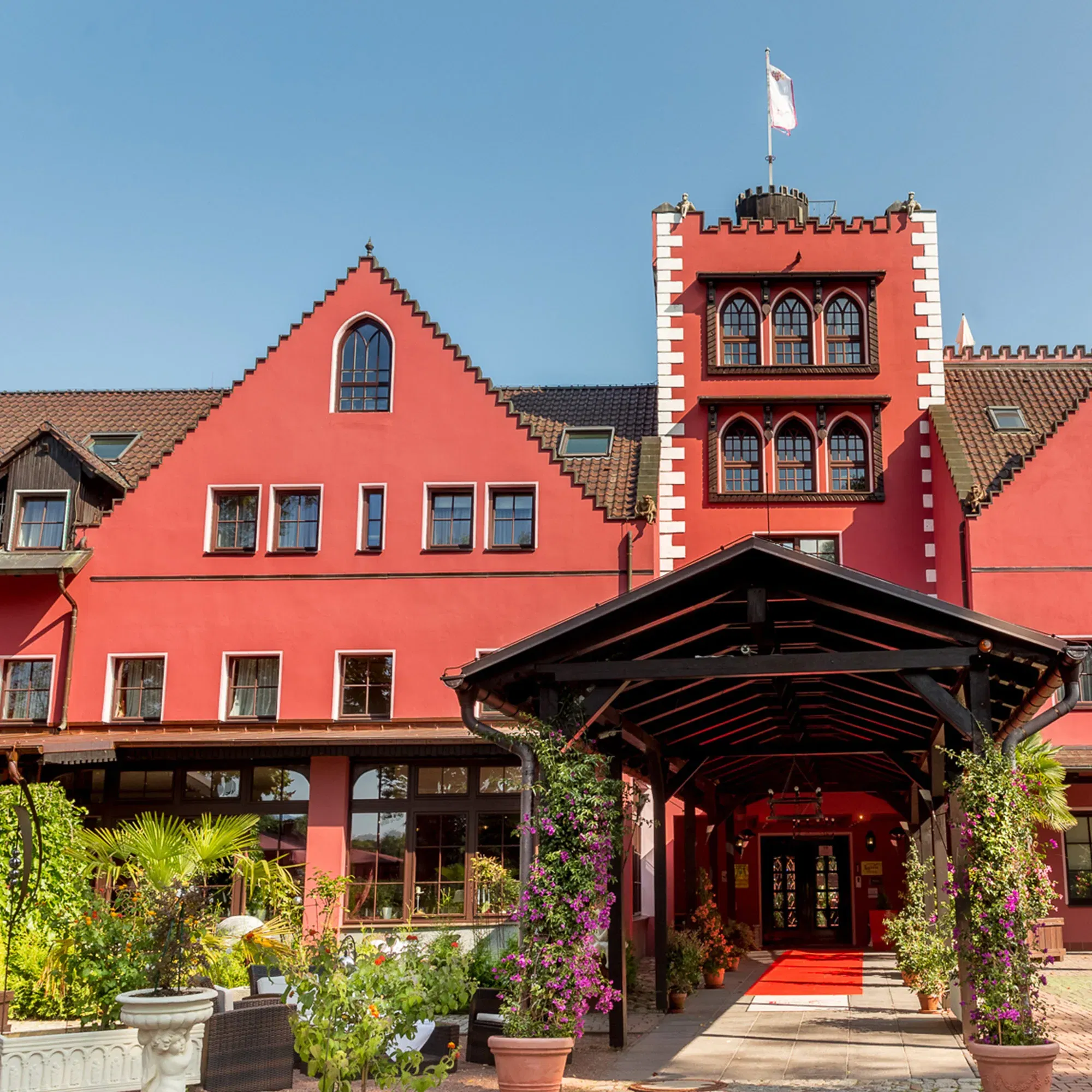 Das Lakeside Hotel, ein markantes Gebäude mit roter Fassade und einem überdachten Eingang, ist umgeben von blühenden Pflanzen. Der rote Teppich führt unter einem blumengeschmückten Baldachin hindurch.