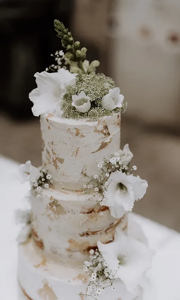 Weiße dreistöckige Hochzeitstorte mit dezenten goldenen Akzenten und Blumen als Dekoration.
