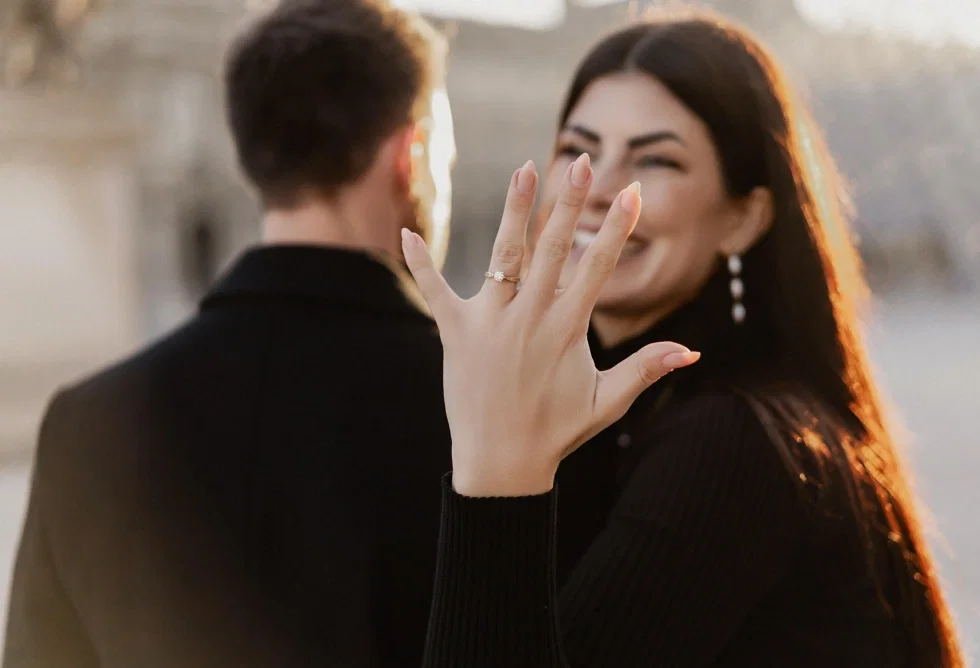 Glückliche Frau zeigt ihren Verlobungsring im Sonnenlicht, während ihr Partner unscharf im Hintergrund steht