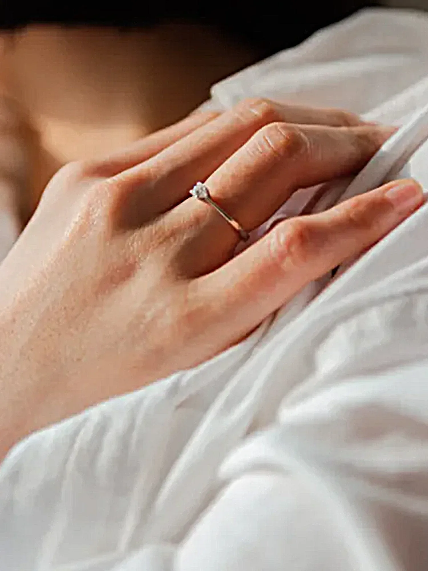 Eine Nahaufnahme einer Hand mit einem schlichten Verlobungsring. Die Hand ruht auf einem weißen Stoff, und der silberne Ring hat einen kleinen, funkelnden Diamanten. Das Bild betont die Eleganz des Rings und den Moment des Verlobungsversprechens.