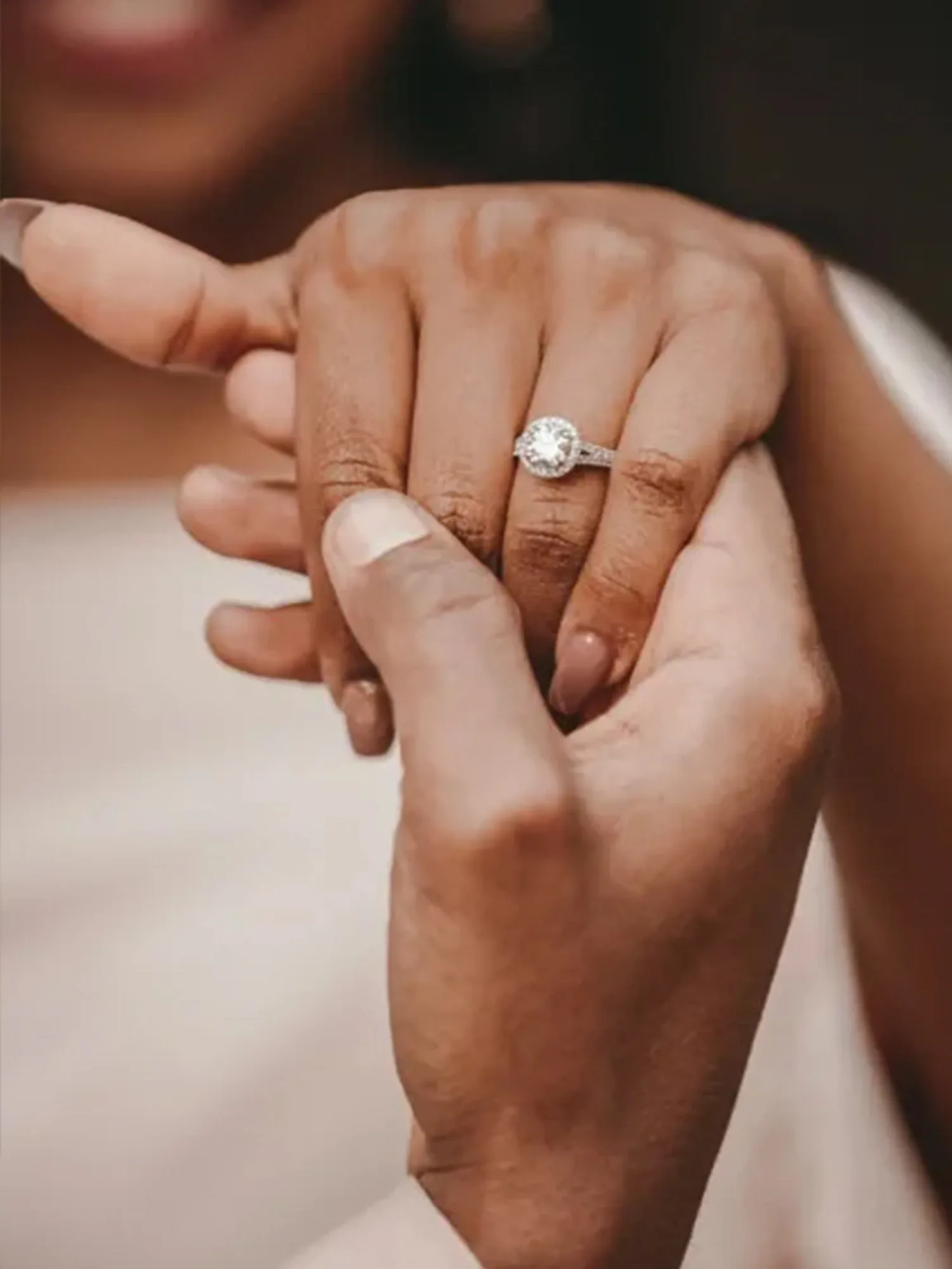 Zwei Hände, eine dunklere und eine hellere, die zusammen einen Finger umschließen, an dem ein Verlobungsring mit einem großen Diamanten steckt. Der Ring ist detailliert zu sehen, während im Hintergrund das glückliche Lächeln der Frau angedeutet wird. Die Geste symbolisiert Nähe und Verbundenheit.