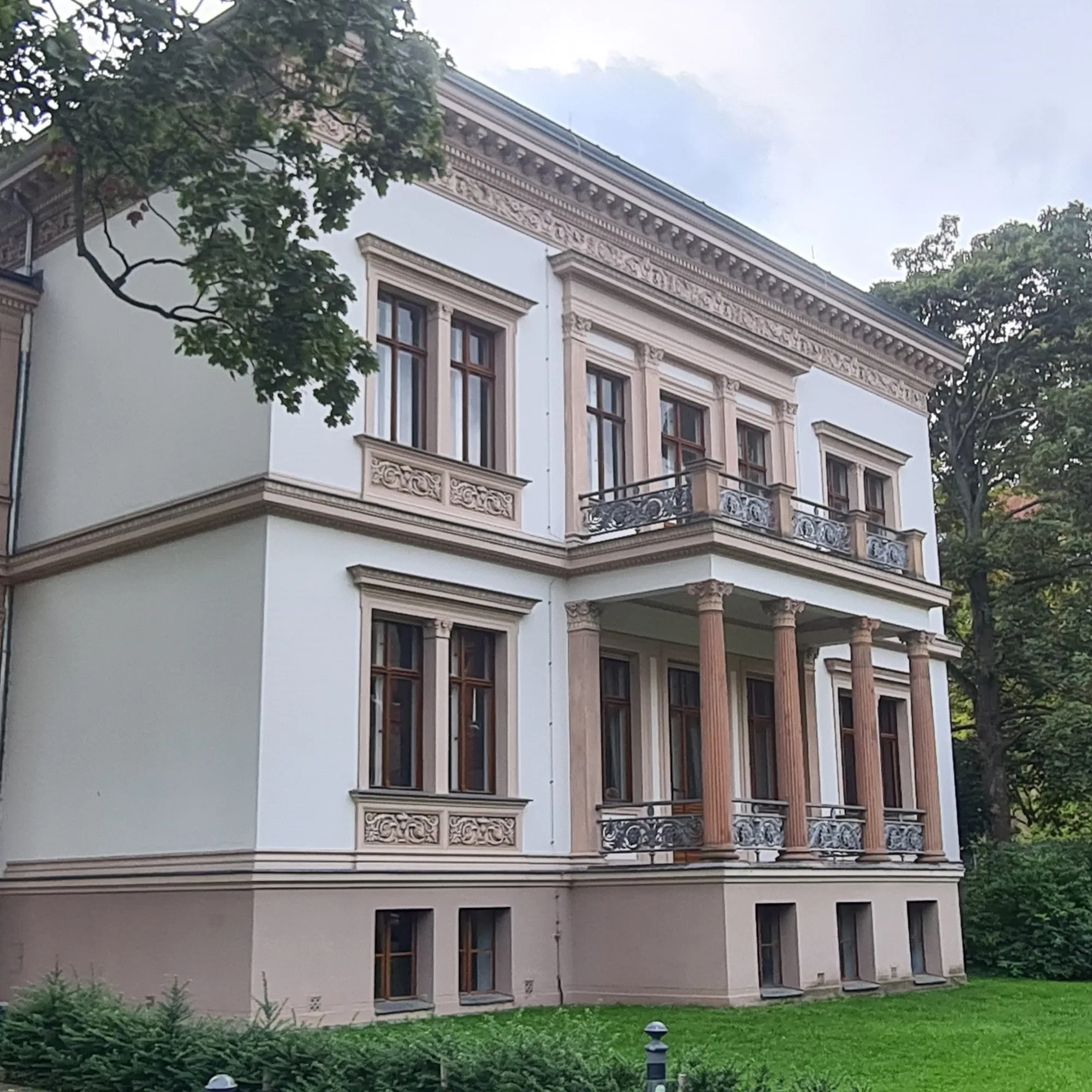 Die Villa Kogge, ein stattliches Gebäude im klassizistischen Stil, mit verzierten Fensterrahmen und zwei symmetrischen Balkonen, die von Säulen getragen werden. Das Gebäude ist von einem grünen Garten umgeben und strahlt Eleganz und Geschichte aus.