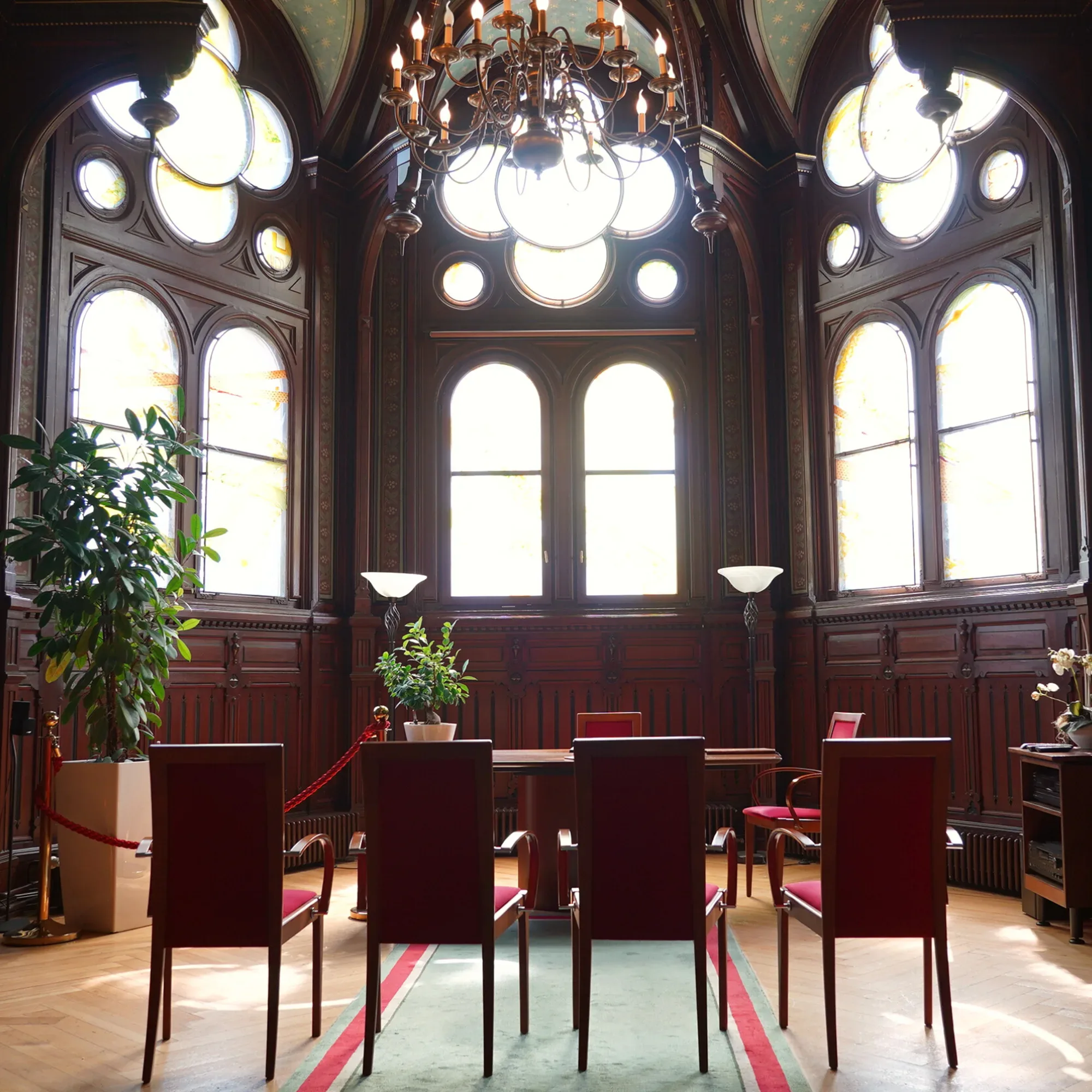 Eine historische Kapelle mit hohen, kunstvoll gestalteten Fenstern, durch die sanftes Licht in den Raum fällt. Der Raum ist mit roten Stühlen und einem großen Kronleuchter ausgestattet, die eine feierliche Atmosphäre schaffen.